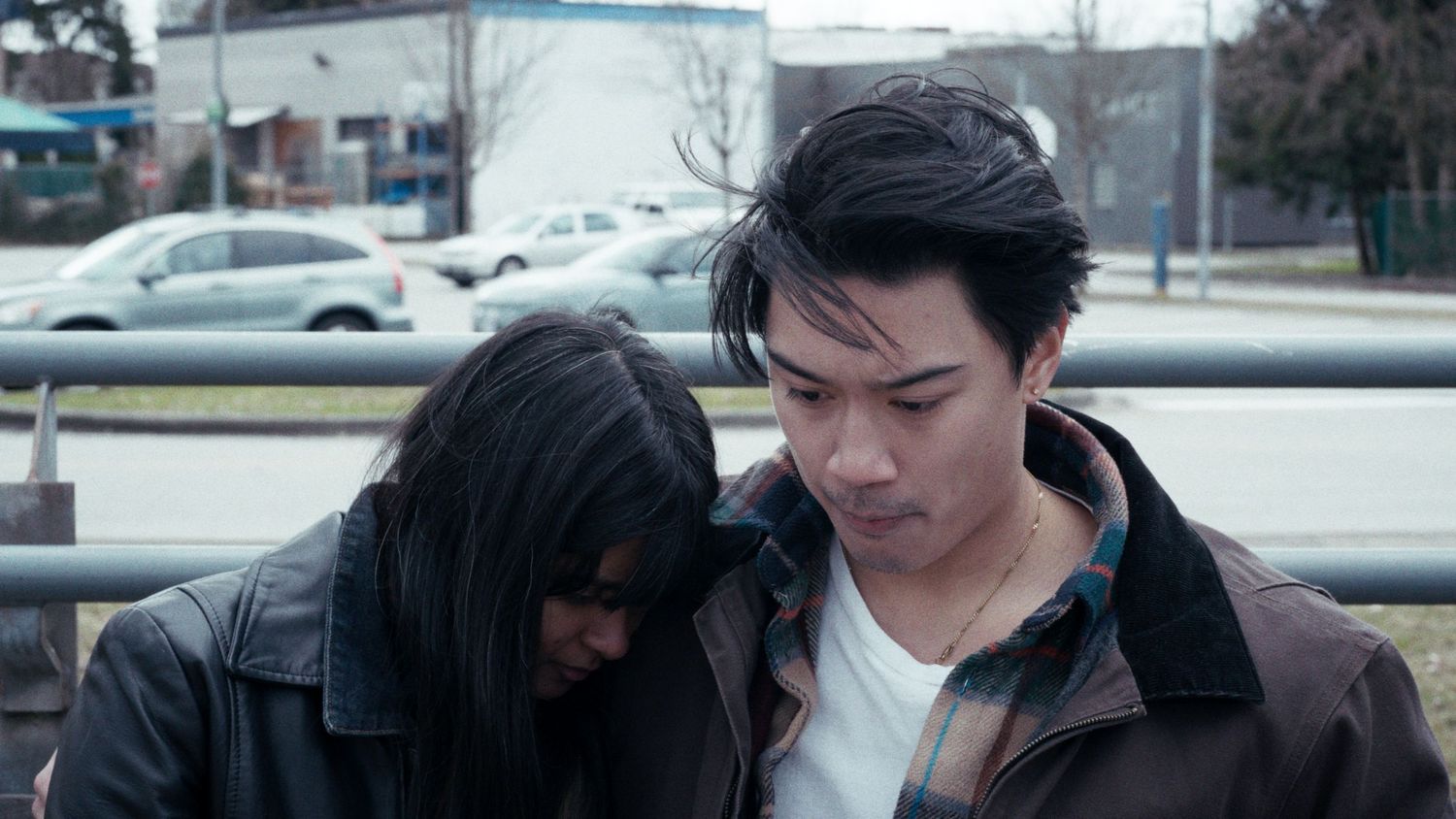 Two young people in leather jackets sitting close together outdoors near a fence.