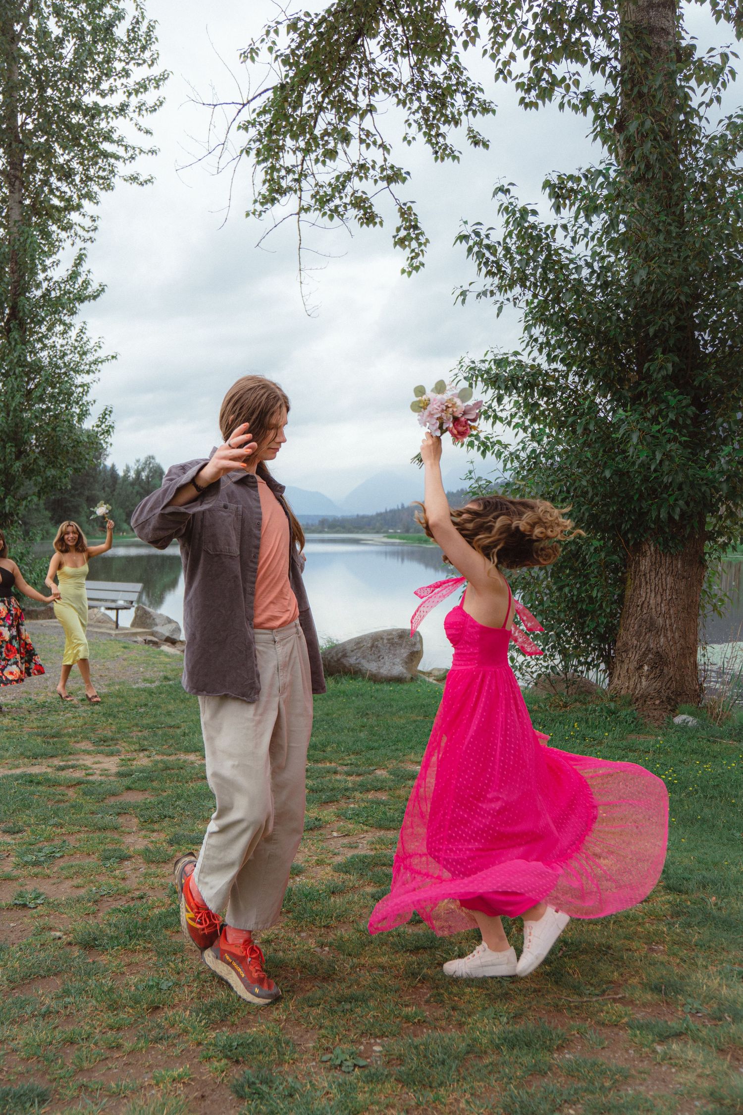 Couple dancing outdoors near lake as woman in flowing pink dress twirls with bouquet.