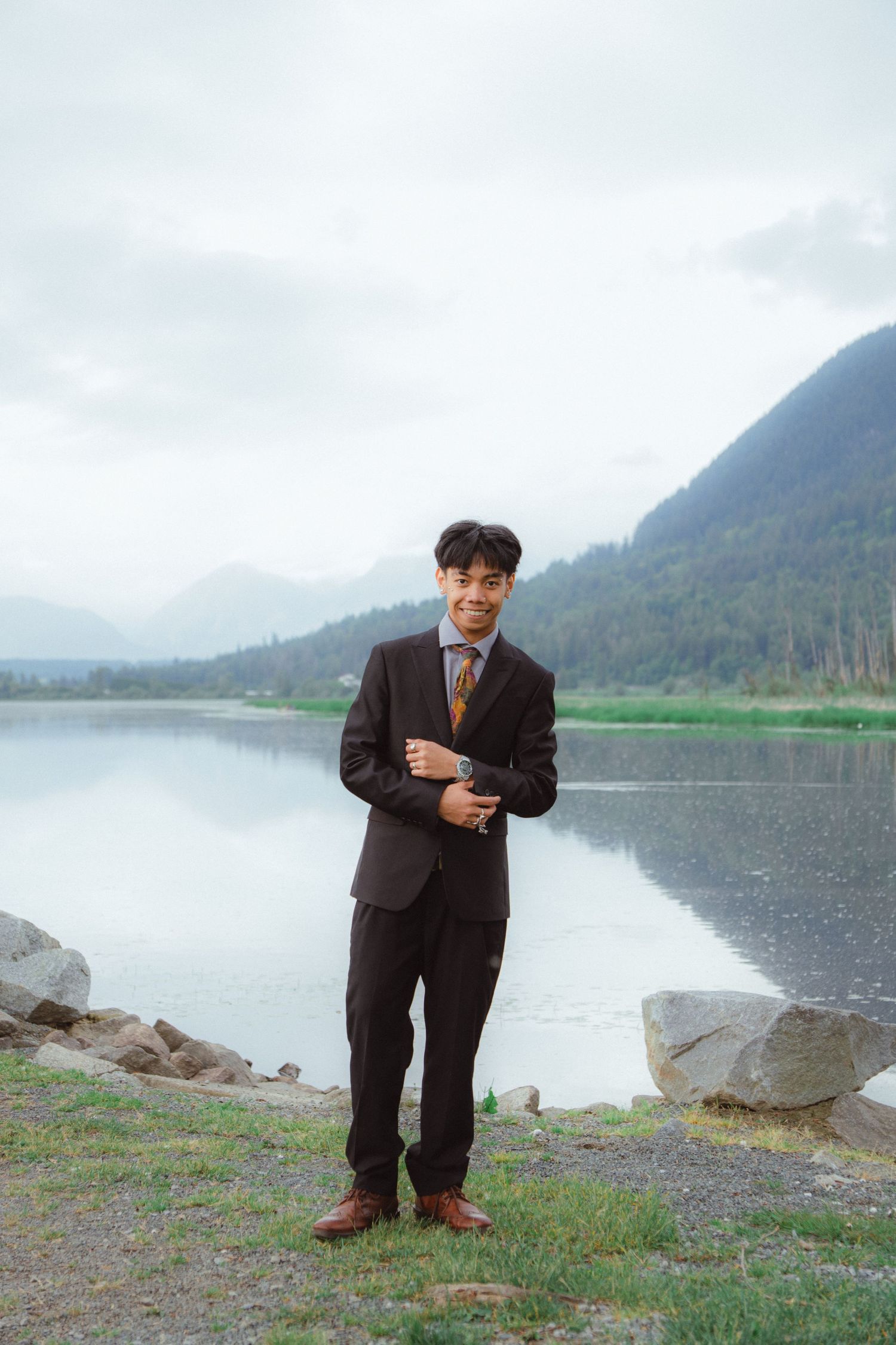 A young man in a dark suit stands confidently by a calm mountain lake.