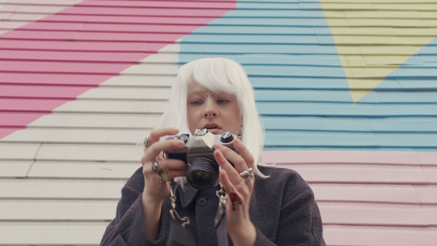 Woman with white hair holding a film camera in front of colorful mural.