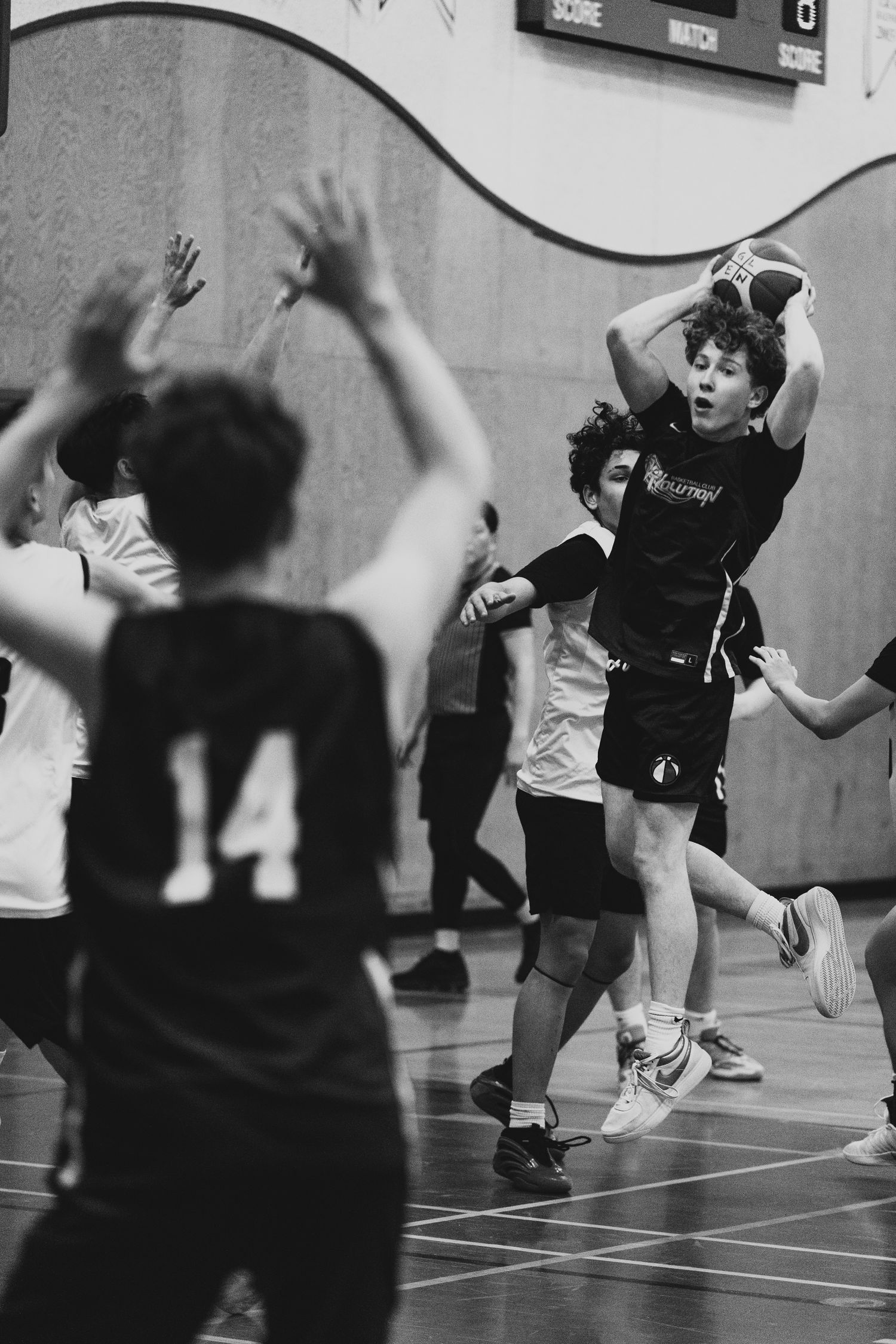 Black and white photo of a young basketball player jumping to pass over defenders in an indoor game.