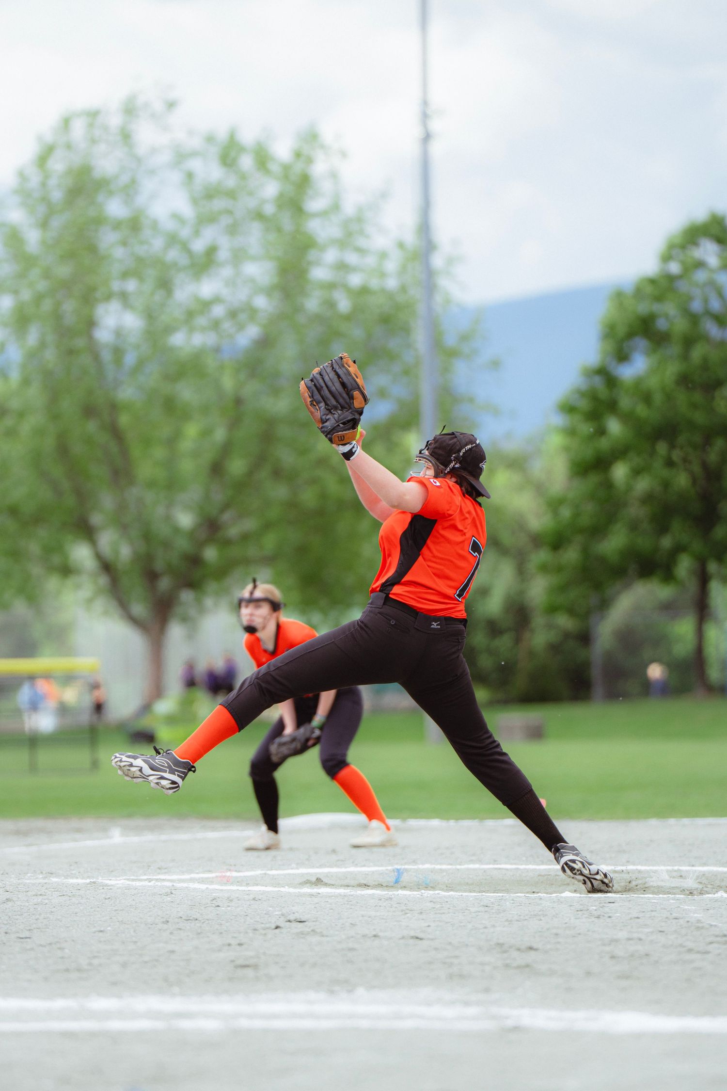 Female softball pitcher in orange and black uniform winds up for a pitch on an outdoor diamond.