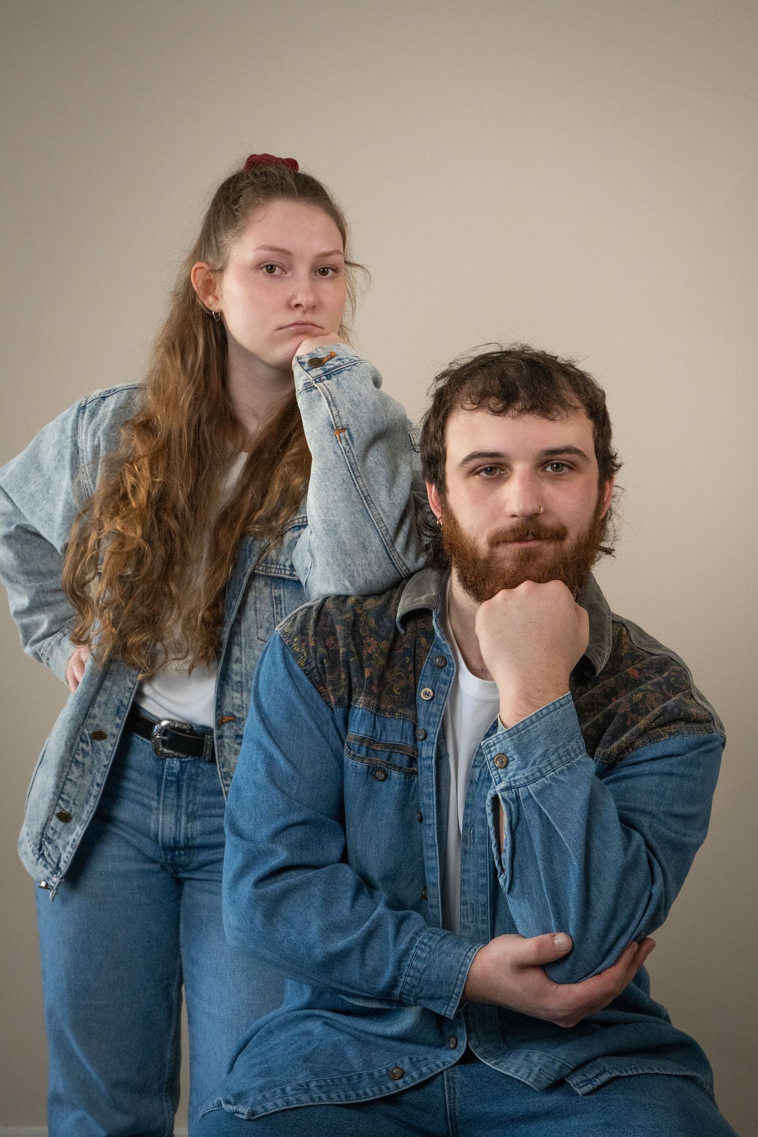 A young woman in a denim jacket rests her hand on a bearded mans shoulder as he sits pensively.