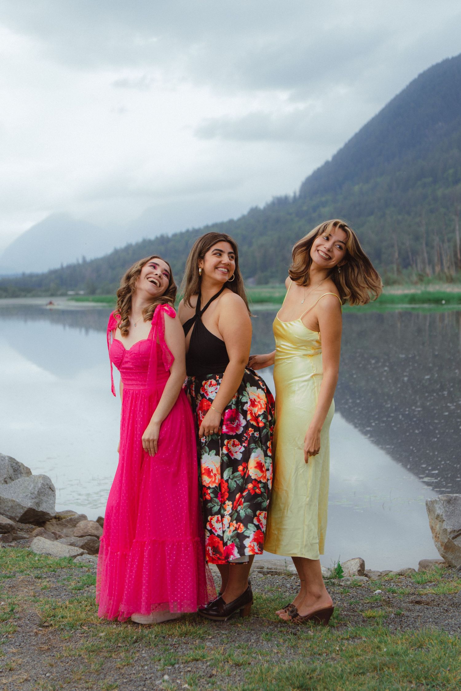 Three laughing women in colorful formal dresses stand together by a scenic mountain lake.