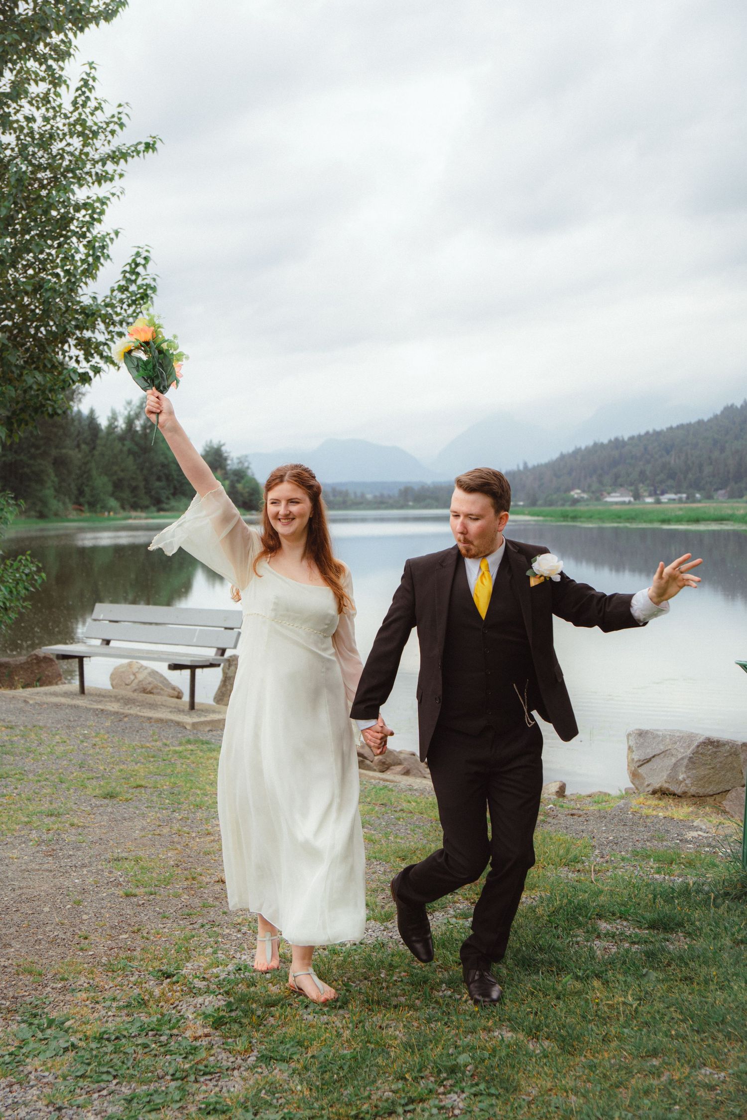 Joyful bride in white dress raising yellow bouquet while groom in black suit walks lakeside.