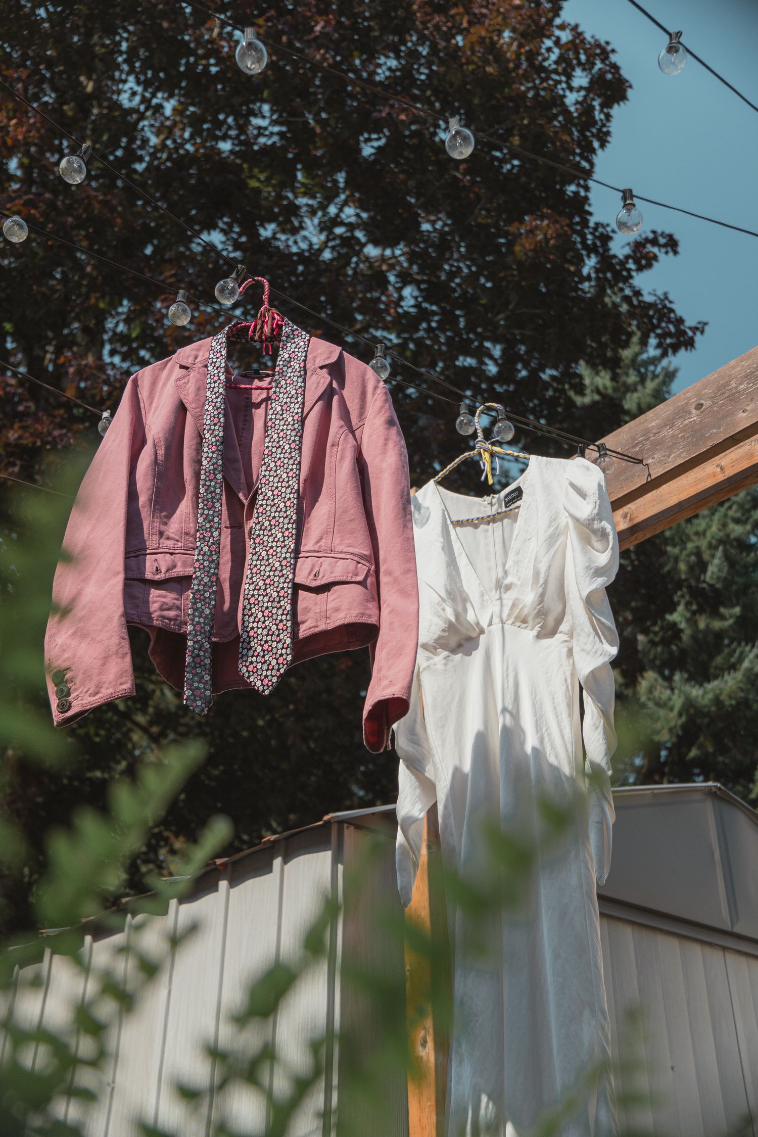 Pink denim jacket with floral tie and white dress hanging on hangers outdoors against a wooden fence.
