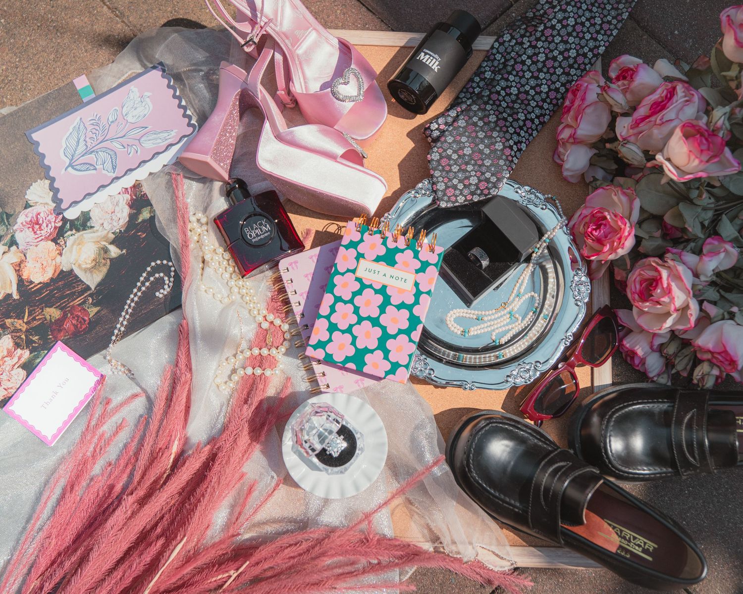 Flat lay of pink roses, black loafers, floral gifts, pearls, and accessories arranged on a surface.