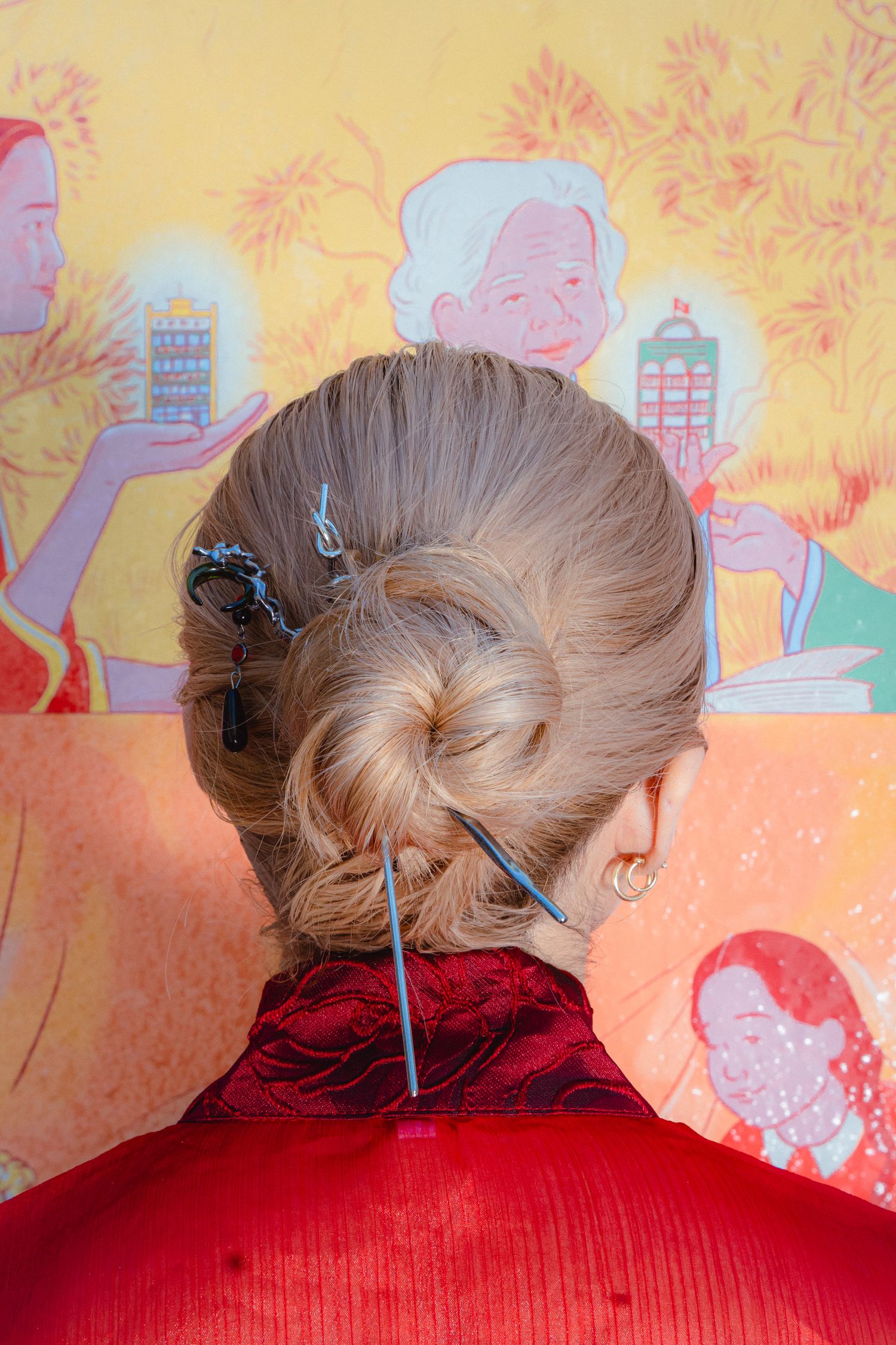 Back view of blonde hair in elegant updo secured with decorative pins, against colorful illustrated mural background.