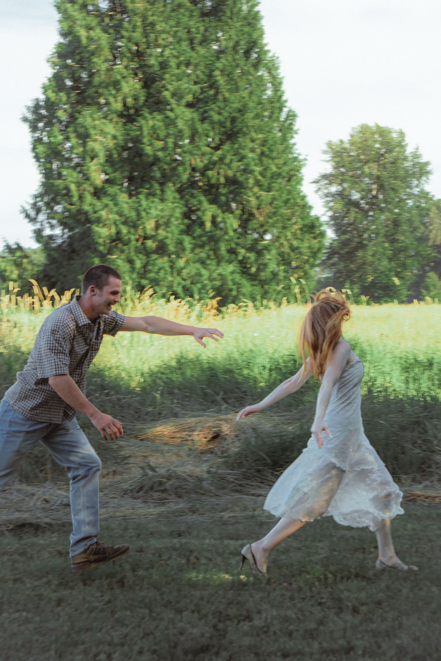 Playful couple running toward each other in a sunny green field, woman in flowing white dress.