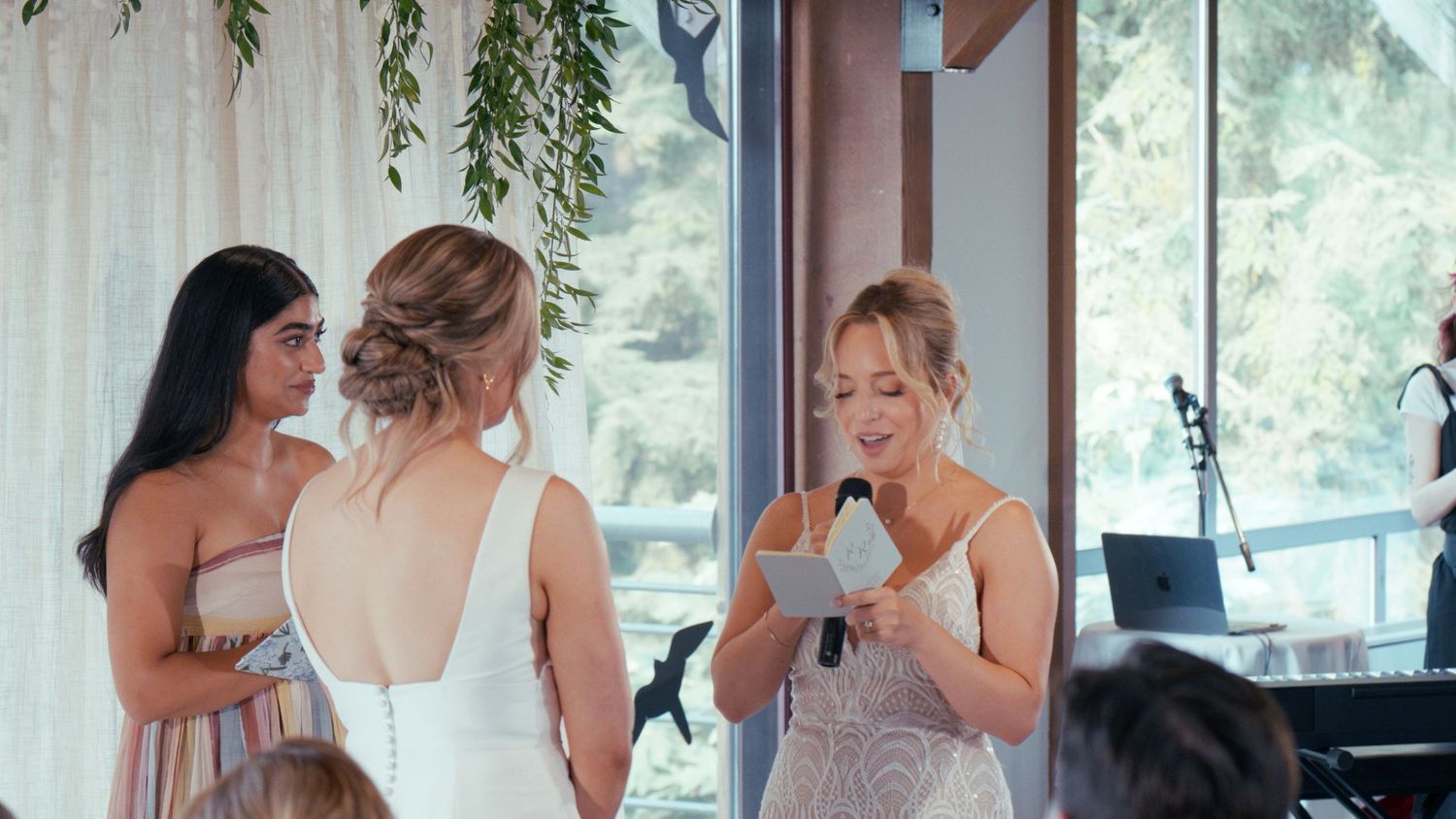 Bride receives heartfelt speech from bridesmaid at elegant indoor wedding ceremony with greenery decor.