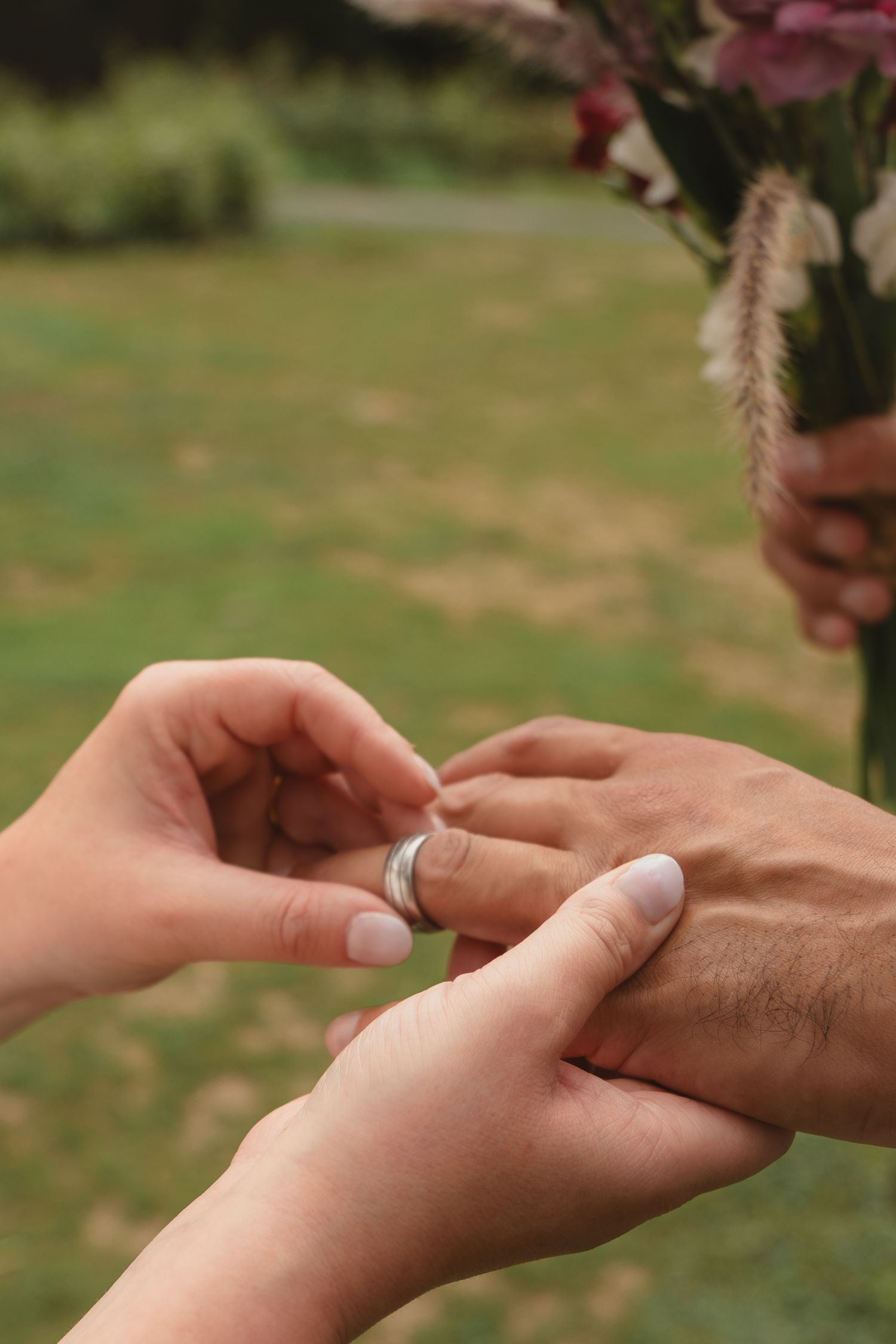 A bride places a silver wedding band on the groom's finger during an outdoor ceremony with green grass background.