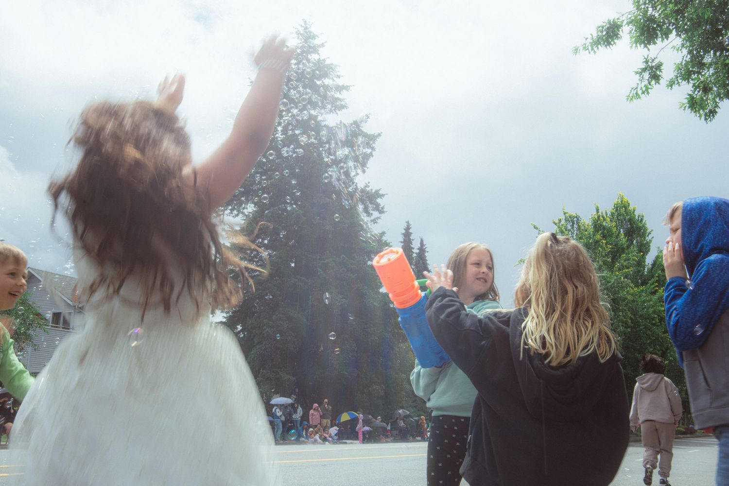 Kids reach for bubbles blown by a bubble gun during an outdoor street parade on a cloudy day.