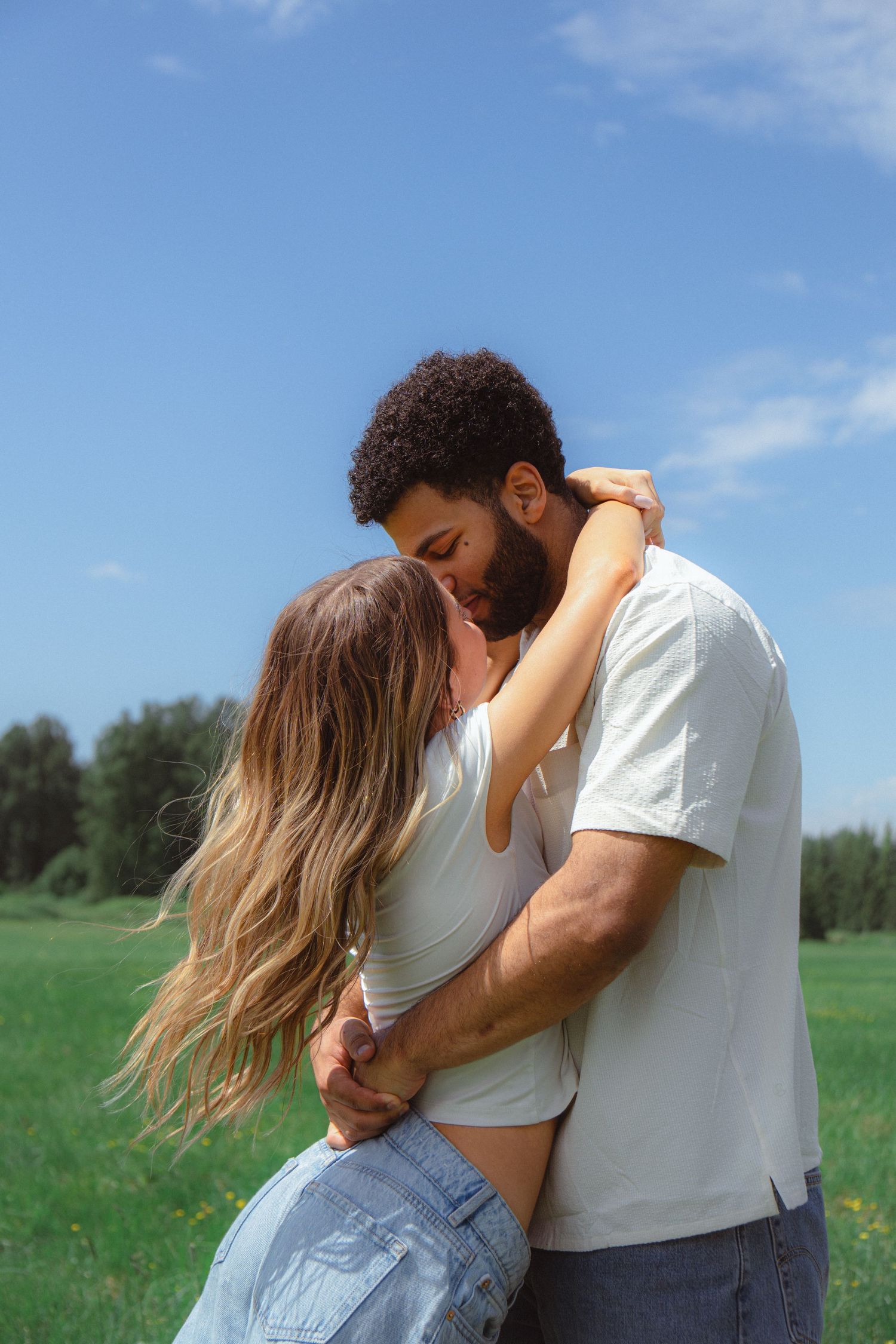 An interracial couple sharing an intimate moment, nearly kissing while embracing in a lush green meadow under blue sky.