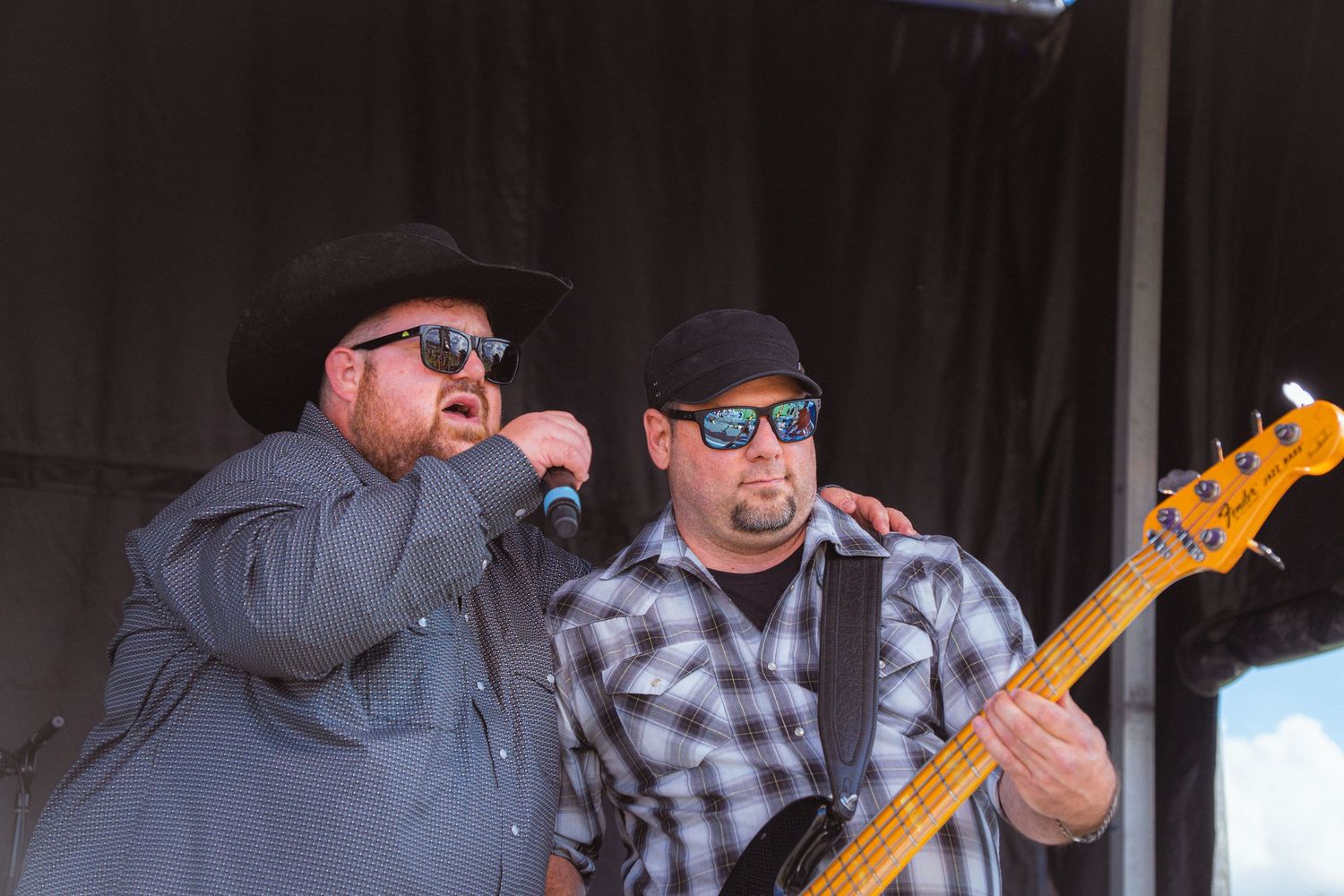 Two men performing on outdoor stage, one singing into mic and one playing bass guitar at festival.