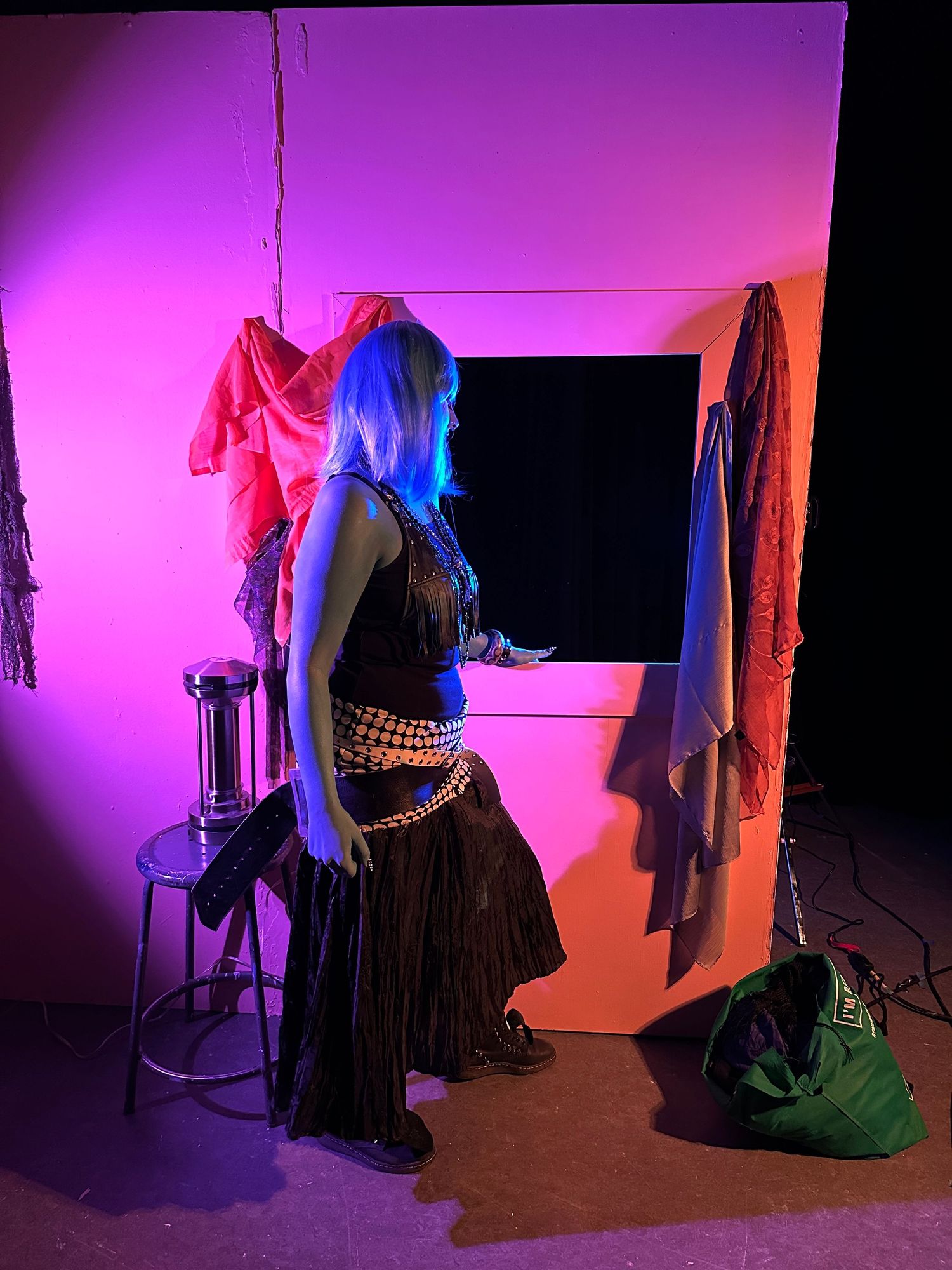 Model with blue hair poses in black outfit against pink and purple neon-lit studio backdrop.
