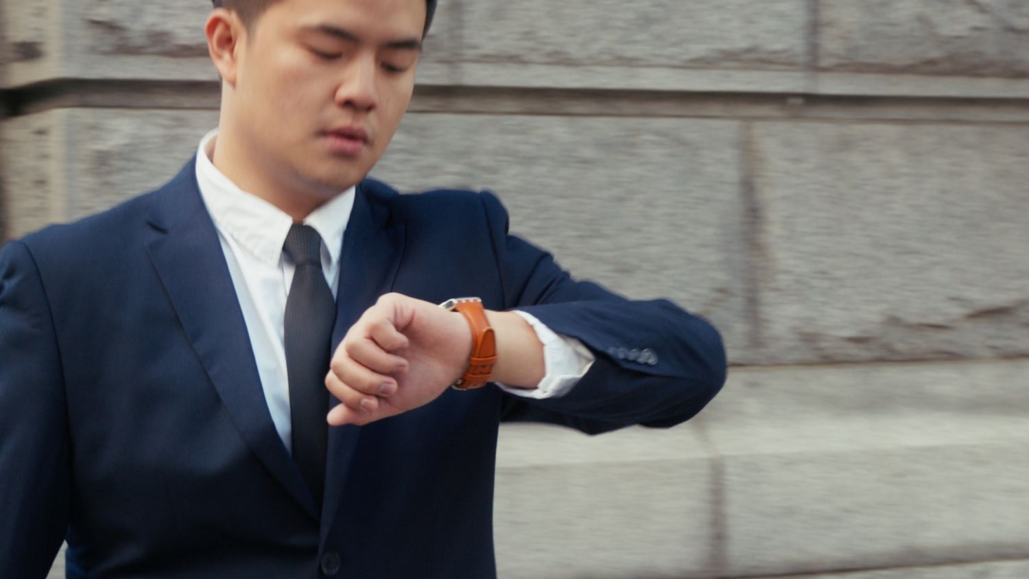Young Asian man in navy blue suit checking time on his brown leather smartwatch outdoors.