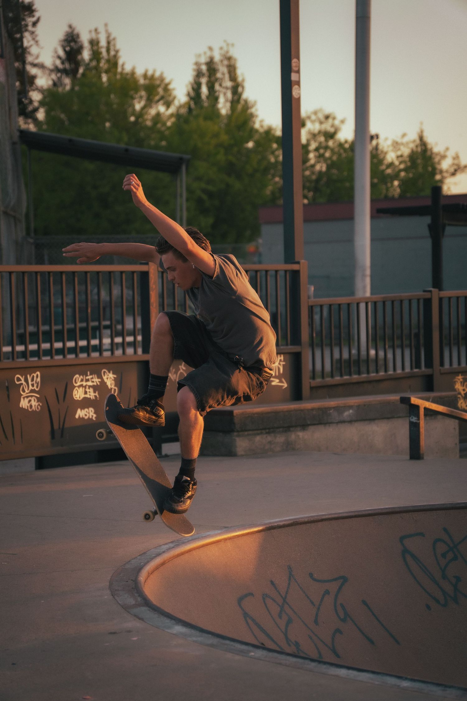 Skateboarder executing an aerial trick above a concrete bowl at a skate park during golden hour.