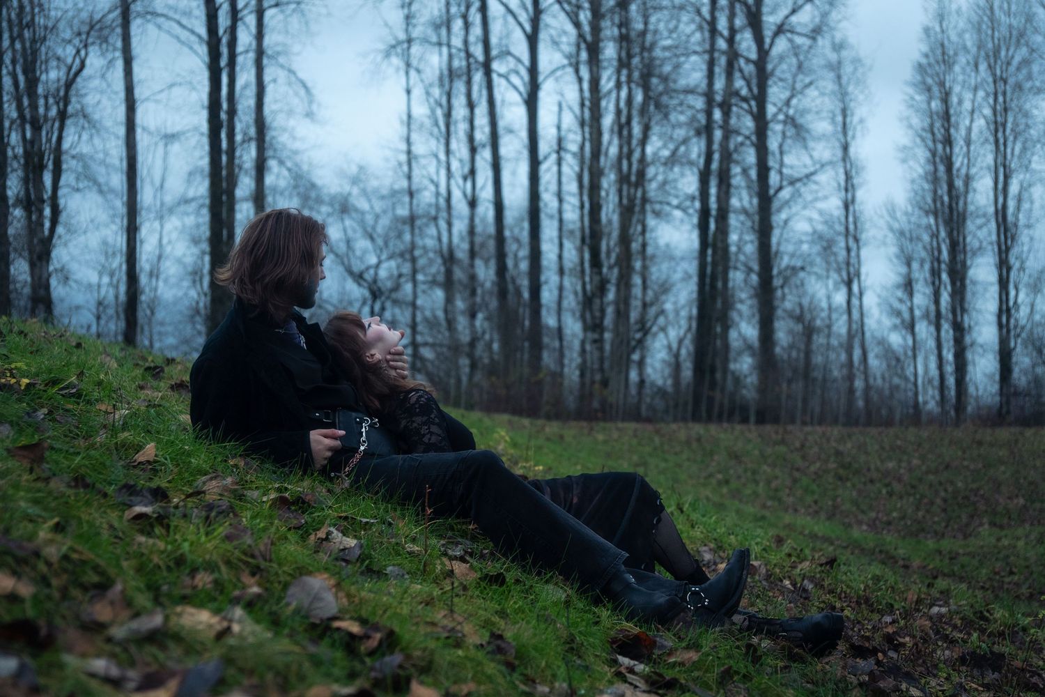 Two people in black sit together on a grassy hillside surrounded by bare trees.