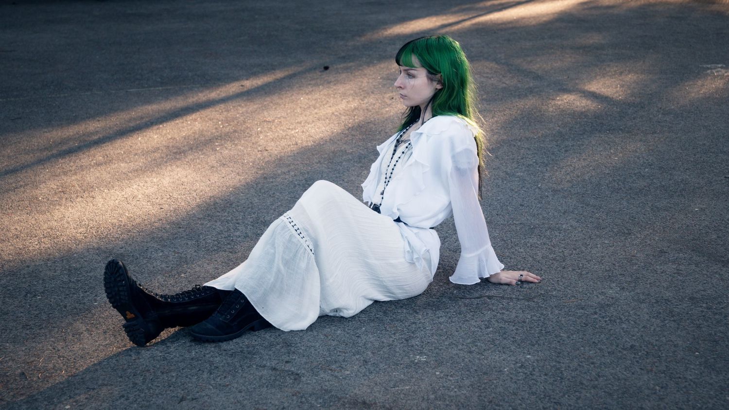 Woman with green hair in white dress and black boots sits relaxed on pavement outdoors.