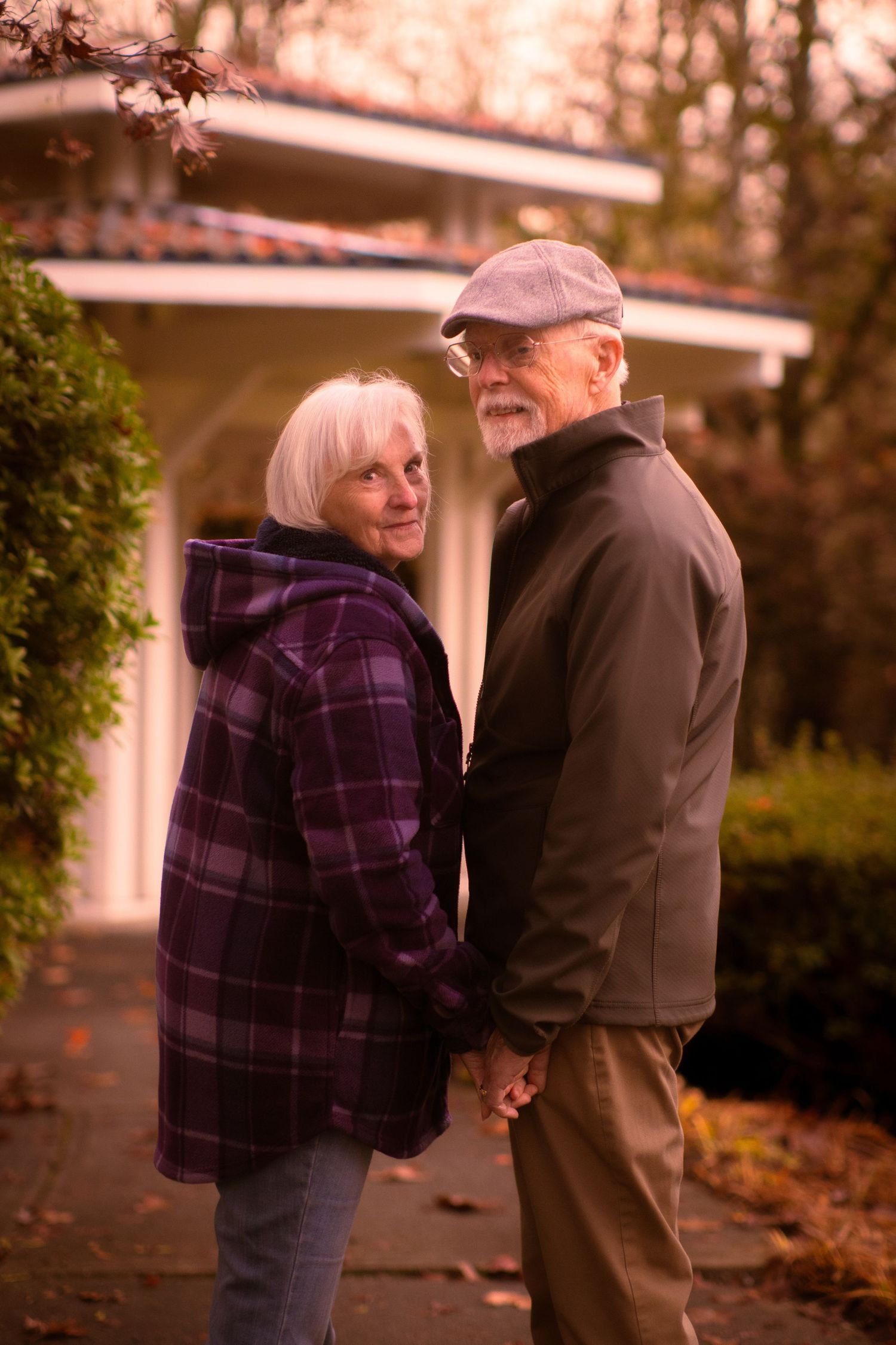 Senior couple holding hands and smiling at each other on a pathway near a home in autumn.