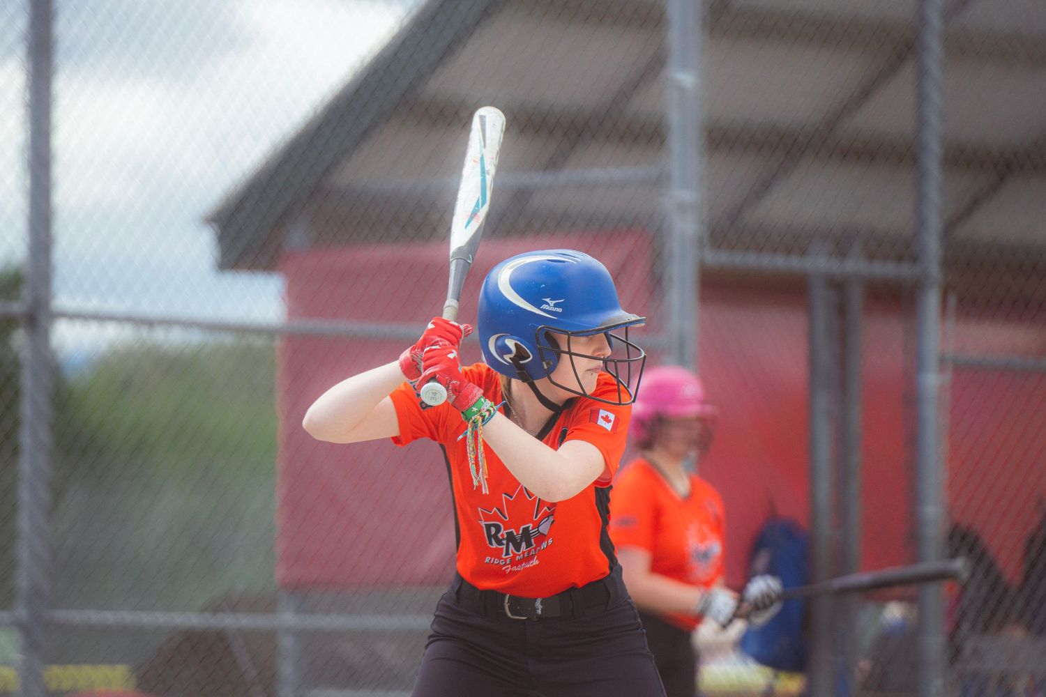 Female softball batter wearing blue helmet and orange RMP jersey prepares to swing.