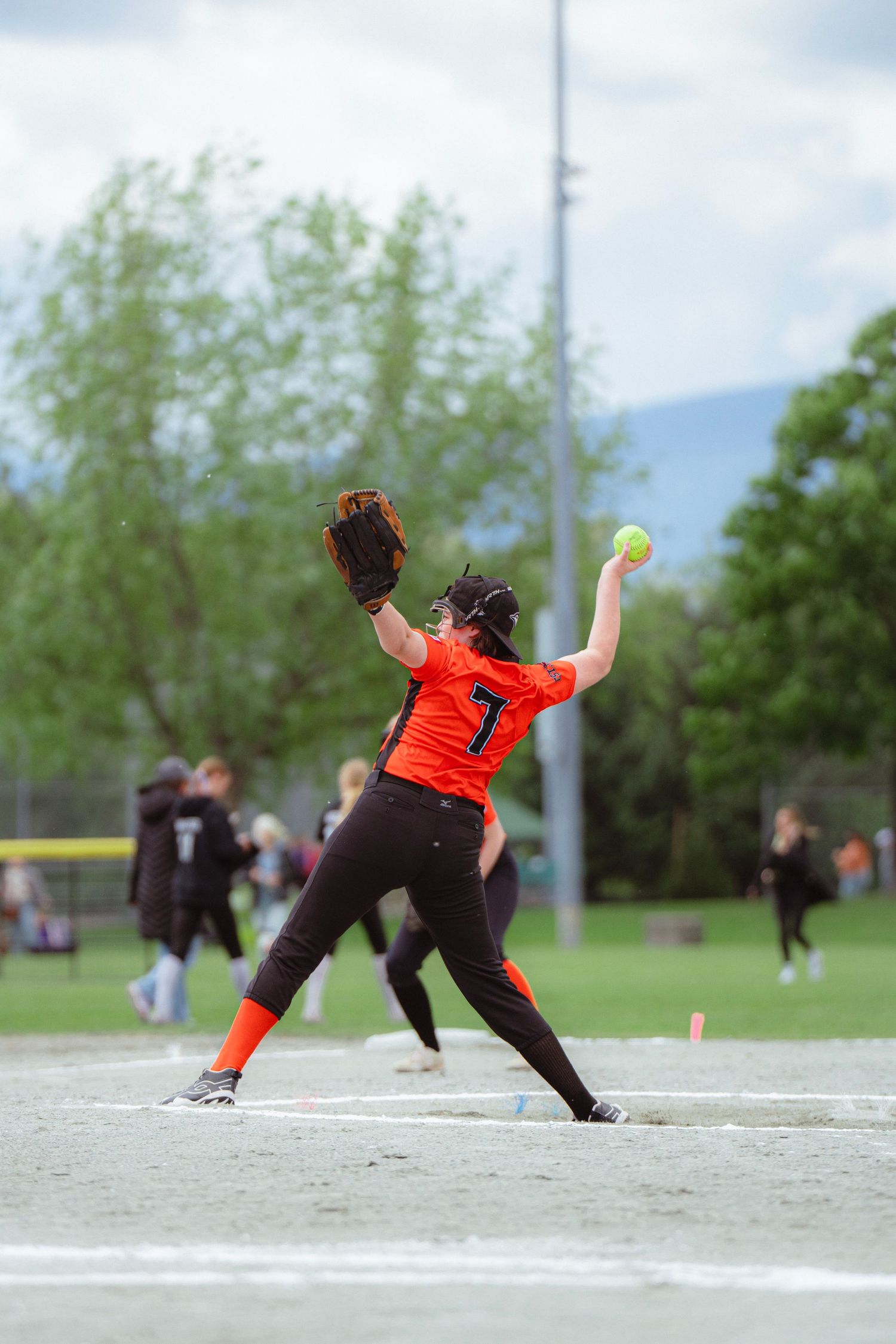 Female softball pitcher wearing number 7 winds up to throw during an outdoor game on a cloudy day.