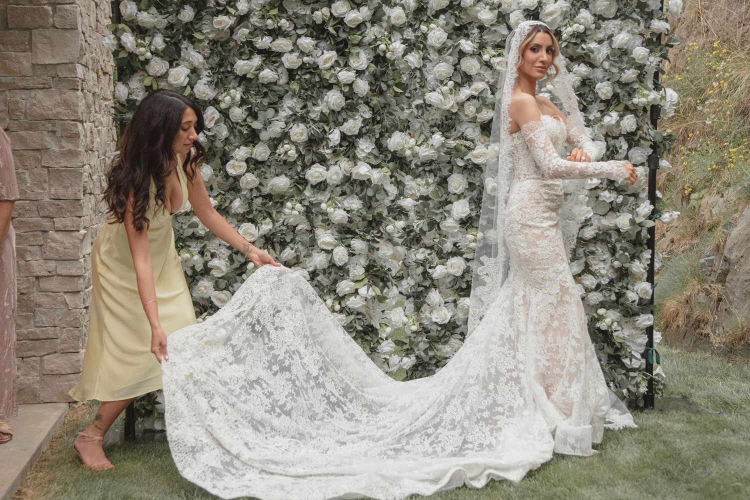 Bride in lace mermaid wedding gown with long train poses before floral wall as bridesmaid arranges dress.