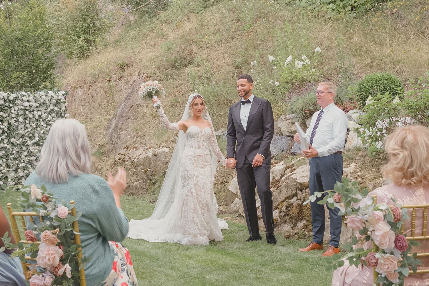 Bride and groom holding hands at outdoor wedding ceremony with floral decorations and lush greenery backdrop.