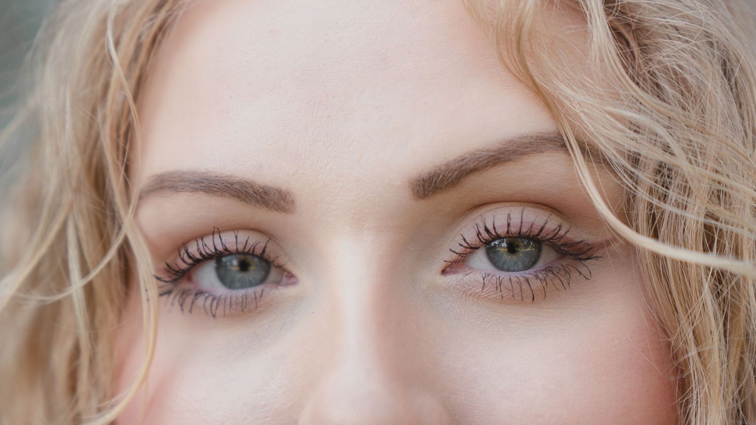 Close-up of a woman with striking blue-green eyes, blonde hair, and long lashes against a soft background.