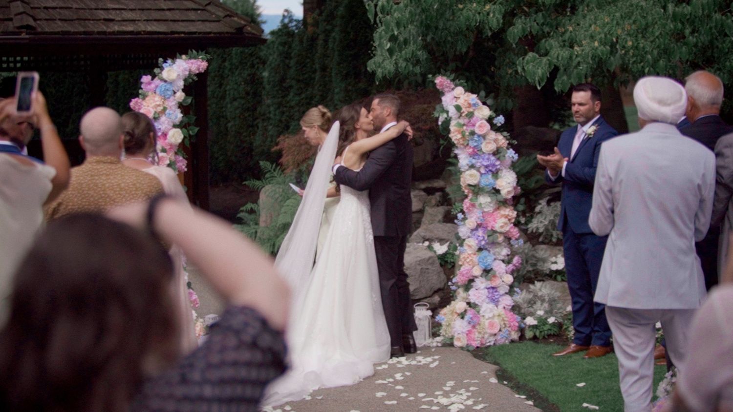 Bride and groom share first kiss at outdoor wedding ceremony with colorful floral arch, surrounded by guests.
