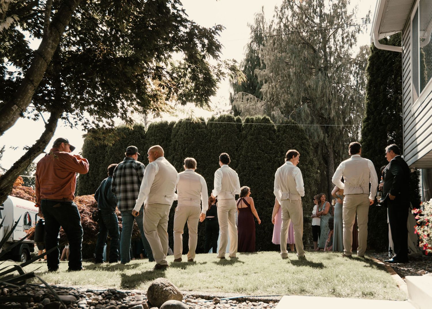 Group of people gathered outdoors in a backyard during a casual event, standing on green grass near trees and hedges.