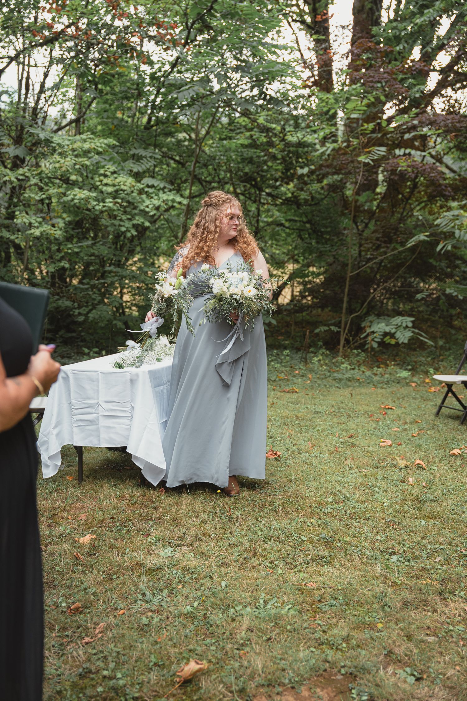 A bridesmaid in a flowing gray gown holds a white floral bouquet outdoors at a garden wedding ceremony.