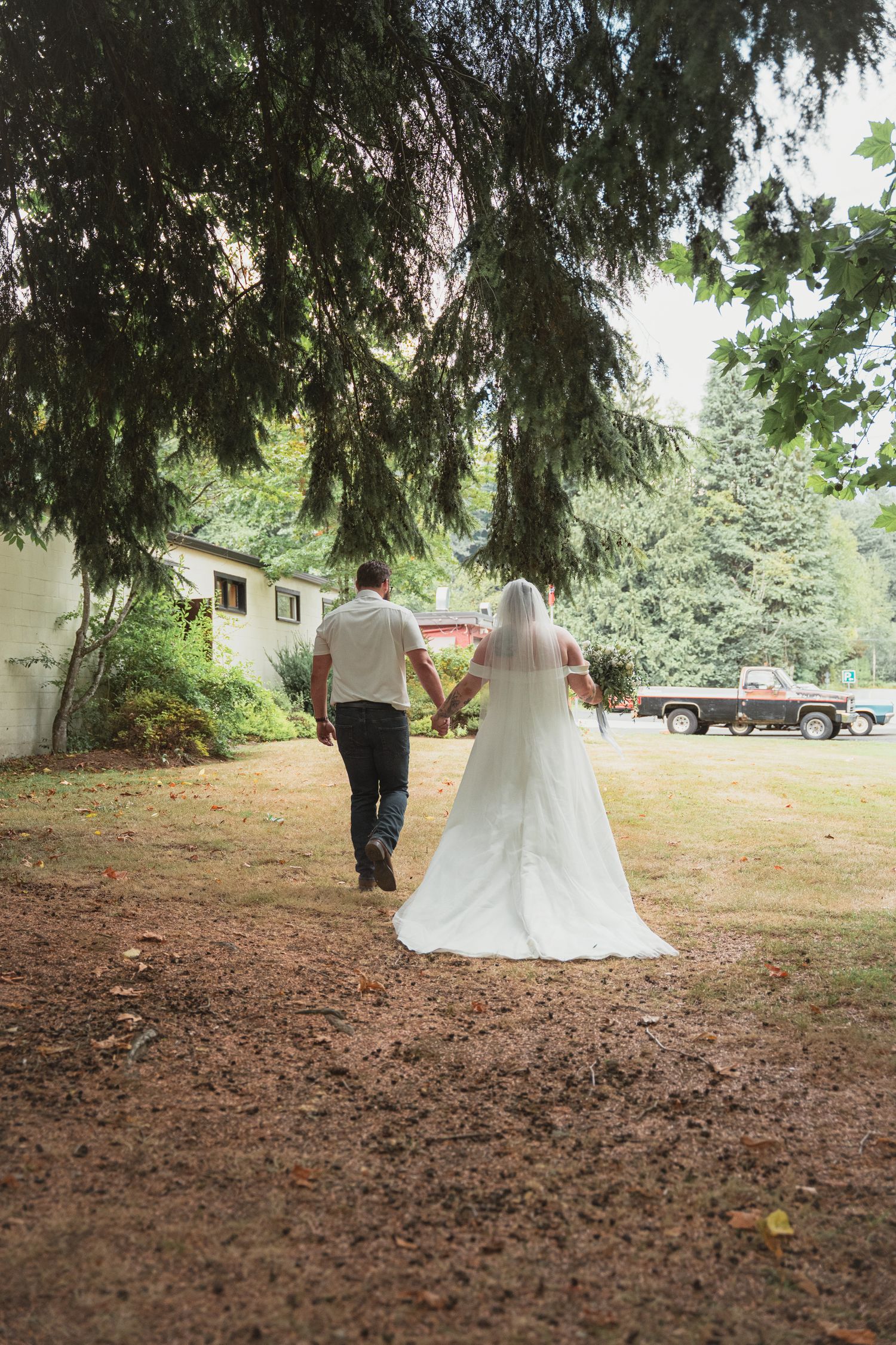 Newlyweds walk hand-in-hand through tree-lined path after outdoor wedding ceremony, bride in white gown with veil.