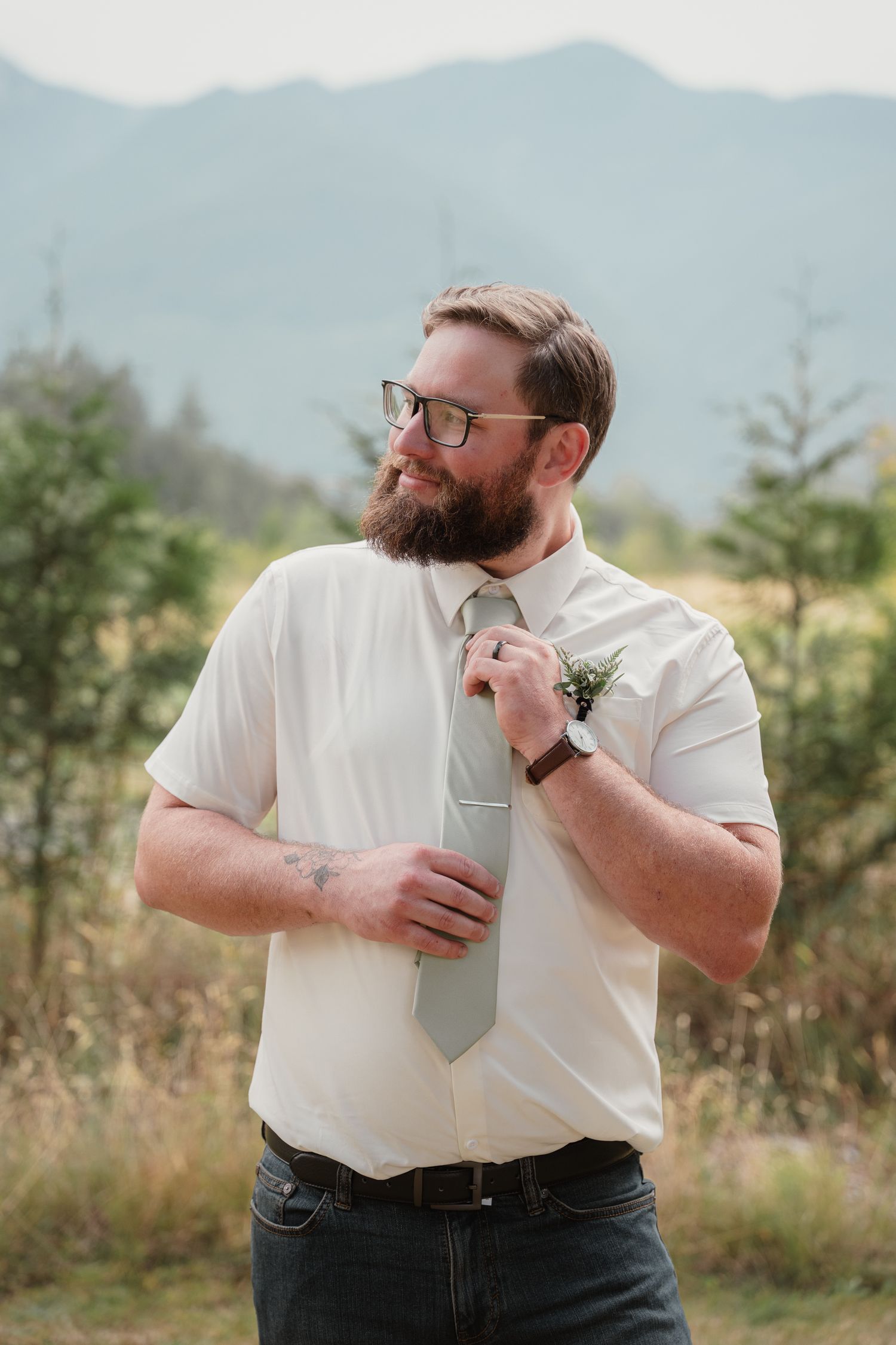 Bearded groom in white shirt adjusting gray tie with boutonniere against mountain backdrop.