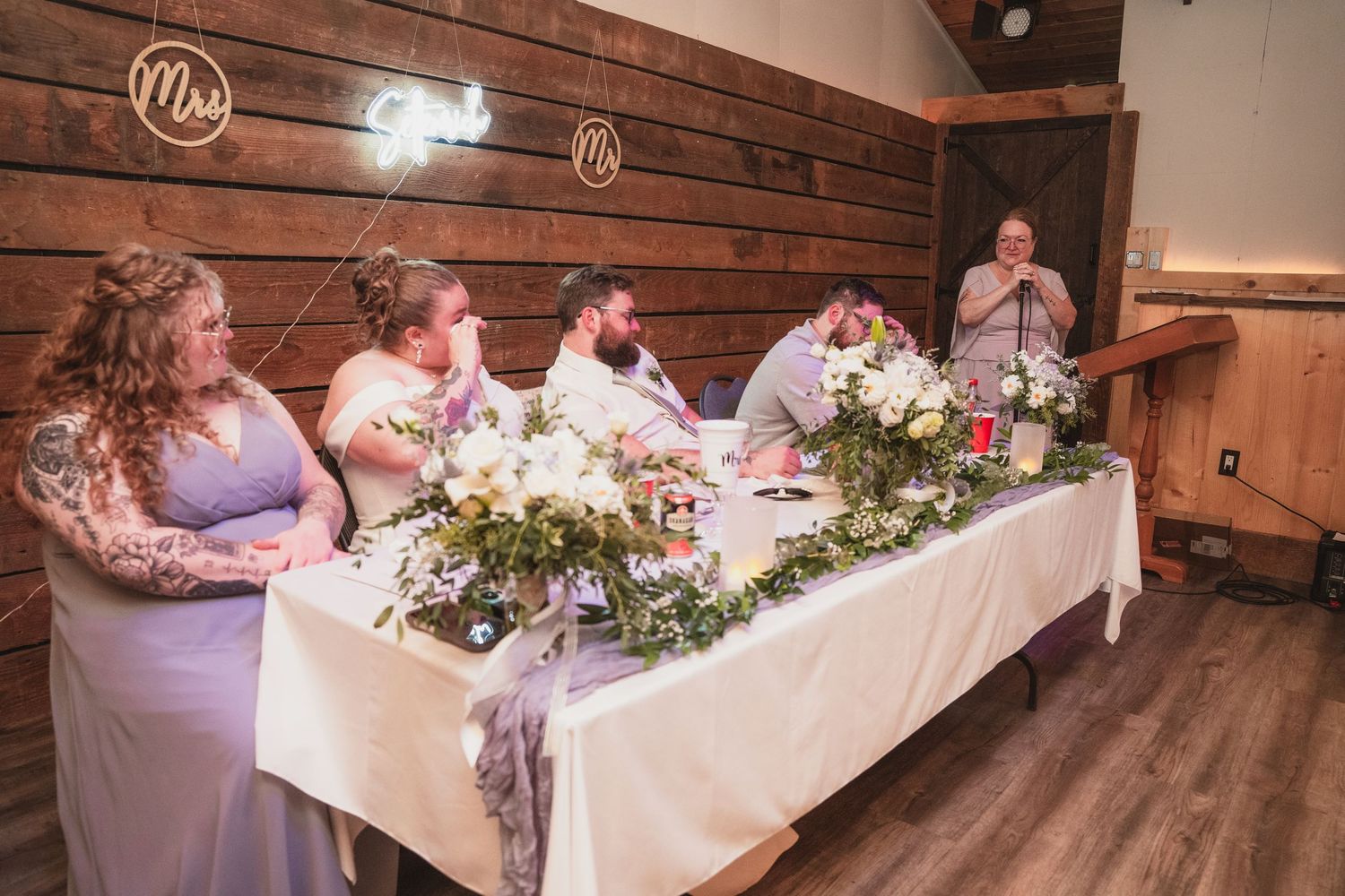 Wedding party at elegantly decorated head table with greenery garland listening to toast in rustic barn reception hall.