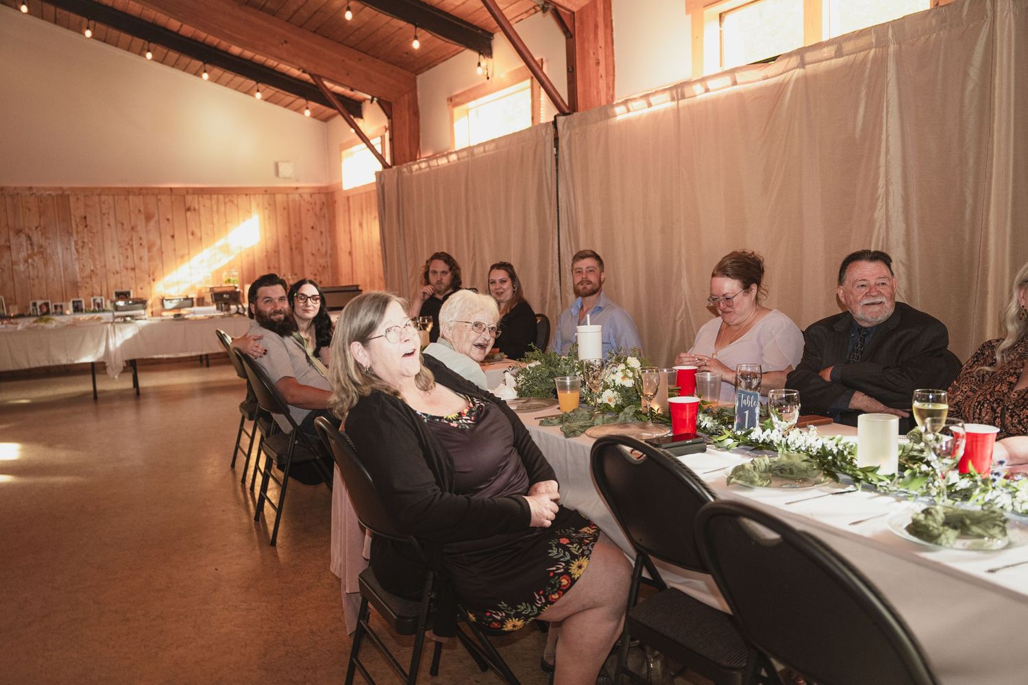 Wedding guests laugh and smile while seated at decorated tables during a rustic barn wedding reception.