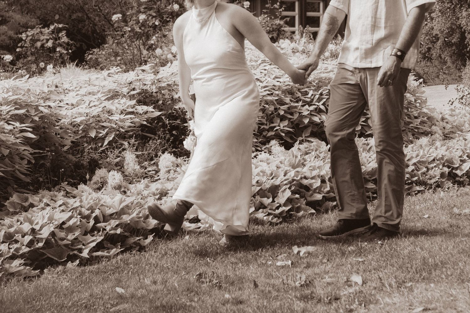 Couple holding hands in a garden, wearing a white wedding dress and casual pants with boots, black and white photo.