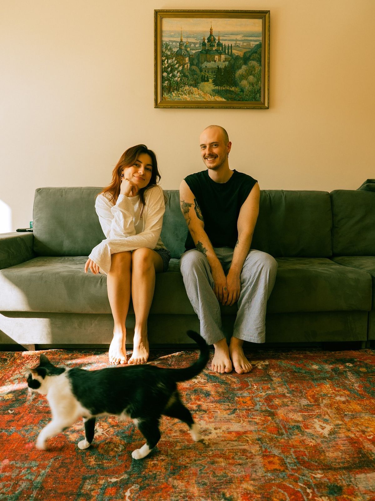 Couple sitting on gray sofa with black and white cat on red rug, painting on wall behind them.