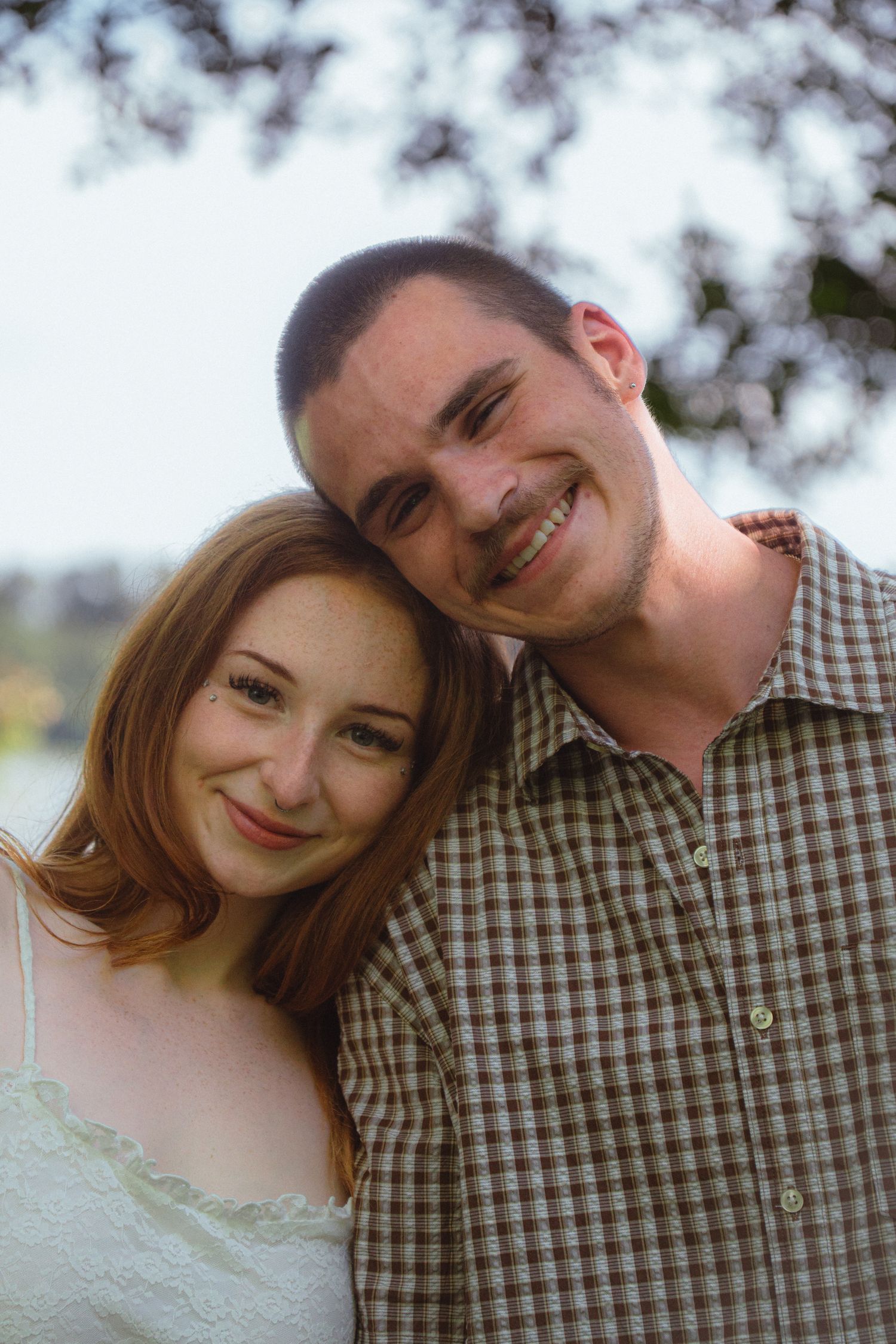 A smiling young couple poses outdoors under blooming trees, the woman leaning on the mans shoulder affectionately.