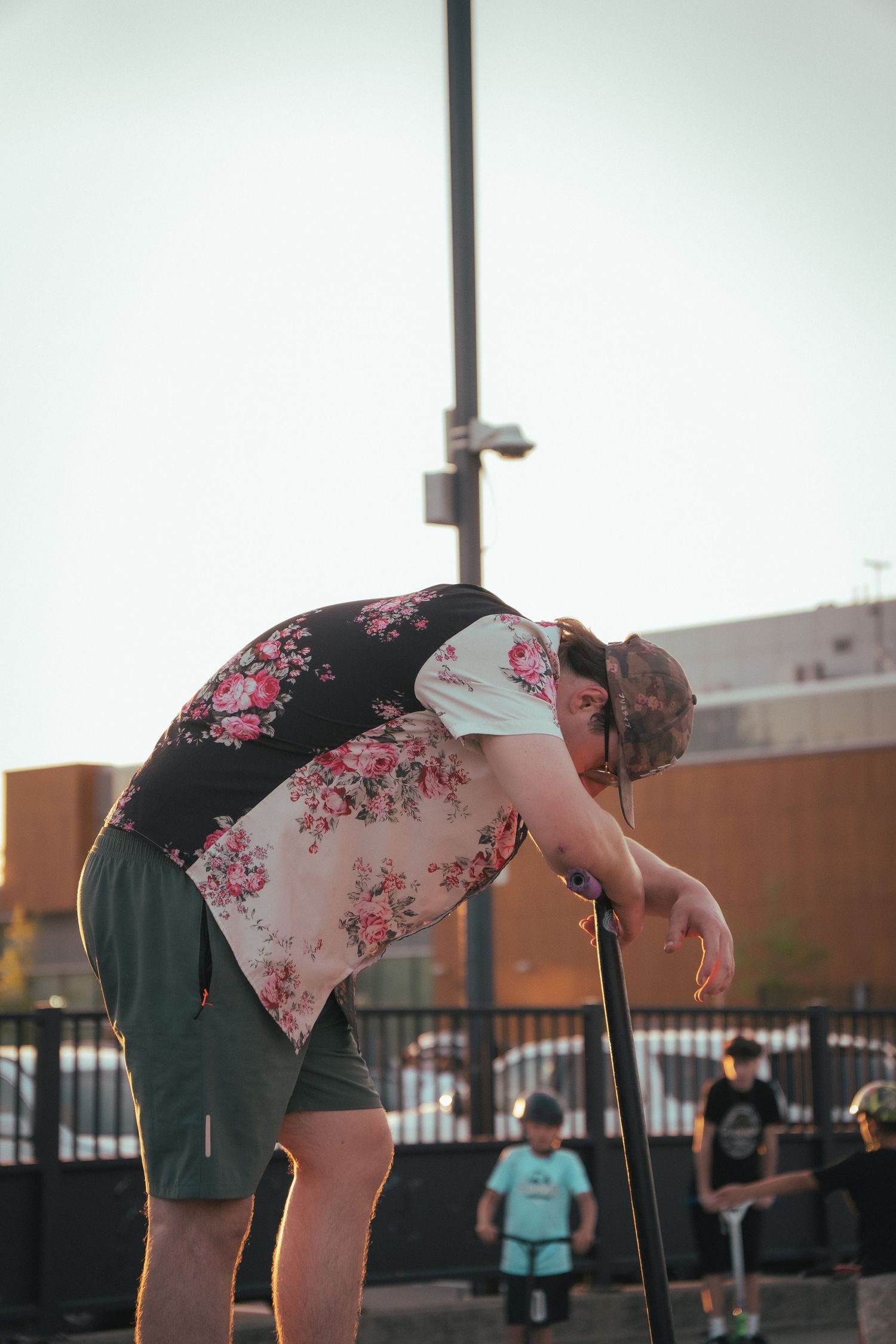Young man in floral shirt bending over a scooter at an outdoor skate park during sunset.