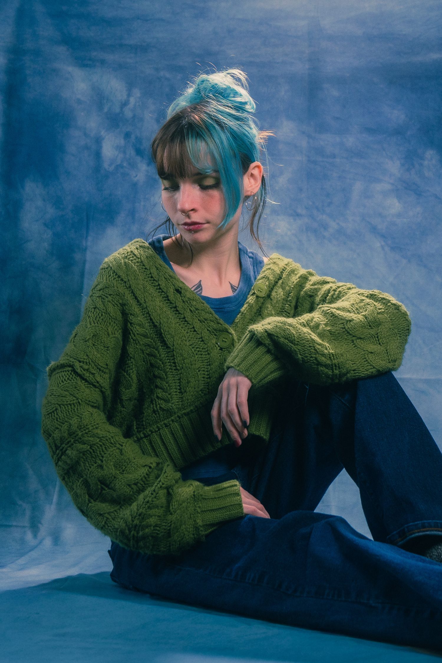 Model with teal updo hairstyle sits pensively in olive cable knit sweater on blue backdrop.