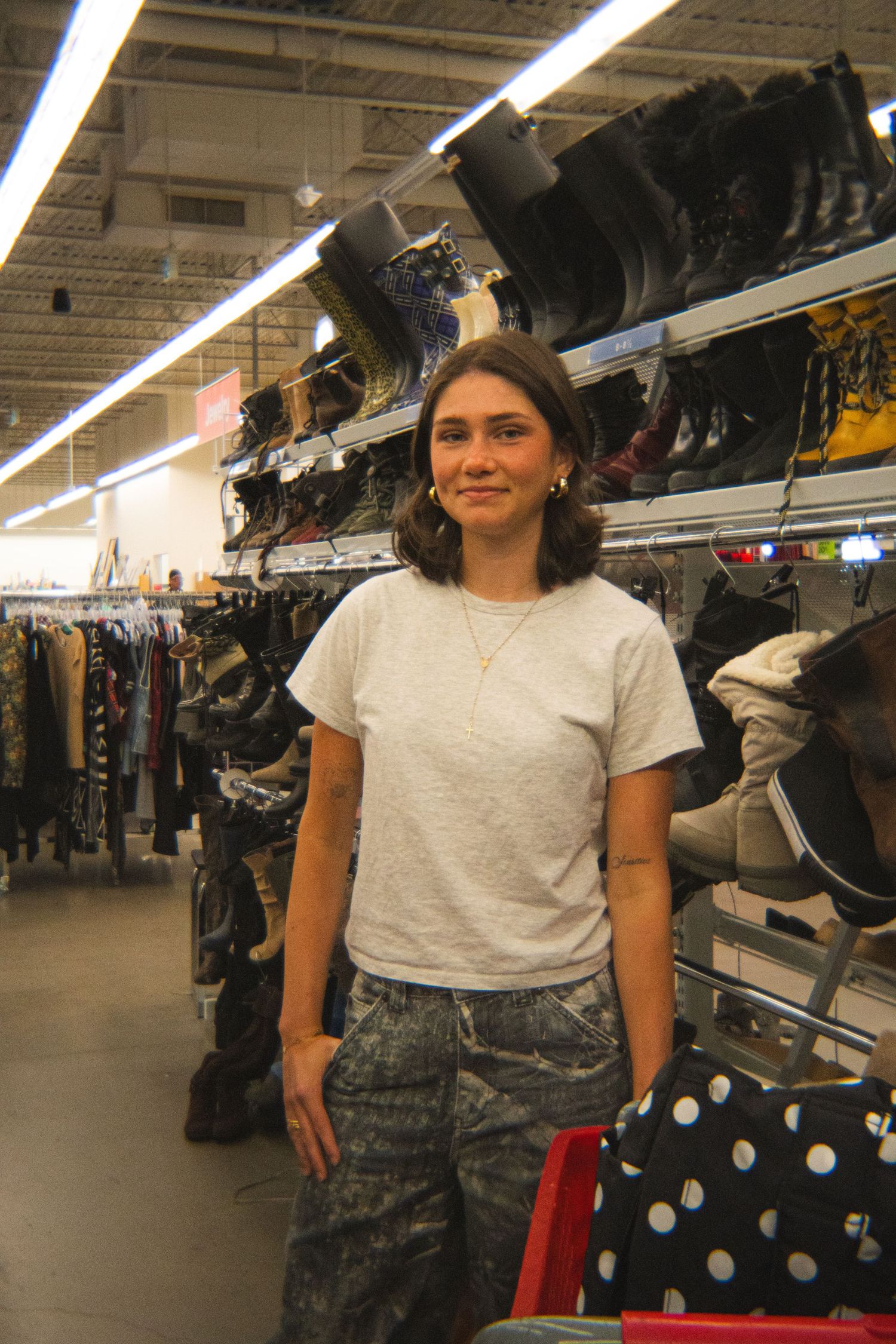 Young woman in gray t-shirt and camo jeans standing in thrift store aisle with shoes and clothing.