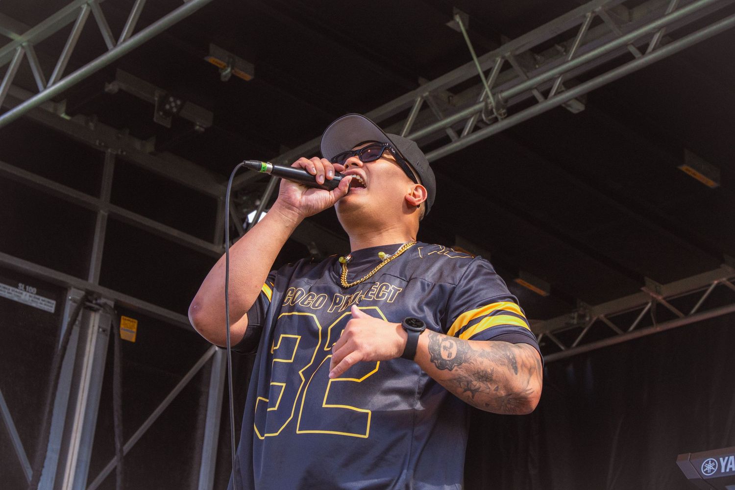 Tattooed male vocalist in number 32 jersey sings passionately into a microphone on outdoor stage.