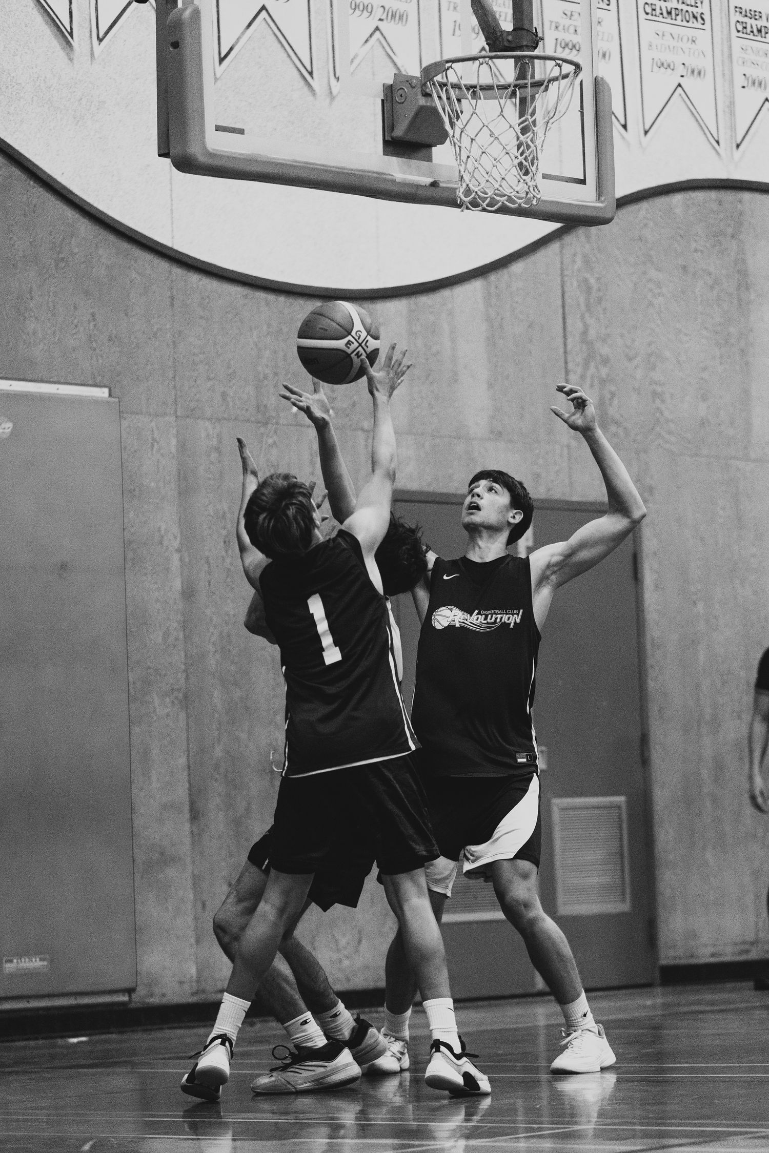Two basketball players in dark jerseys compete for a rebound under the hoop during an indoor game.