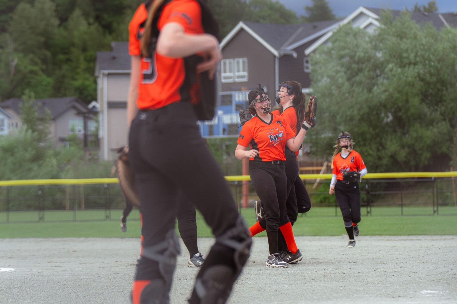 Female softball team in orange uniforms gathered on field during rainy outdoor game day.