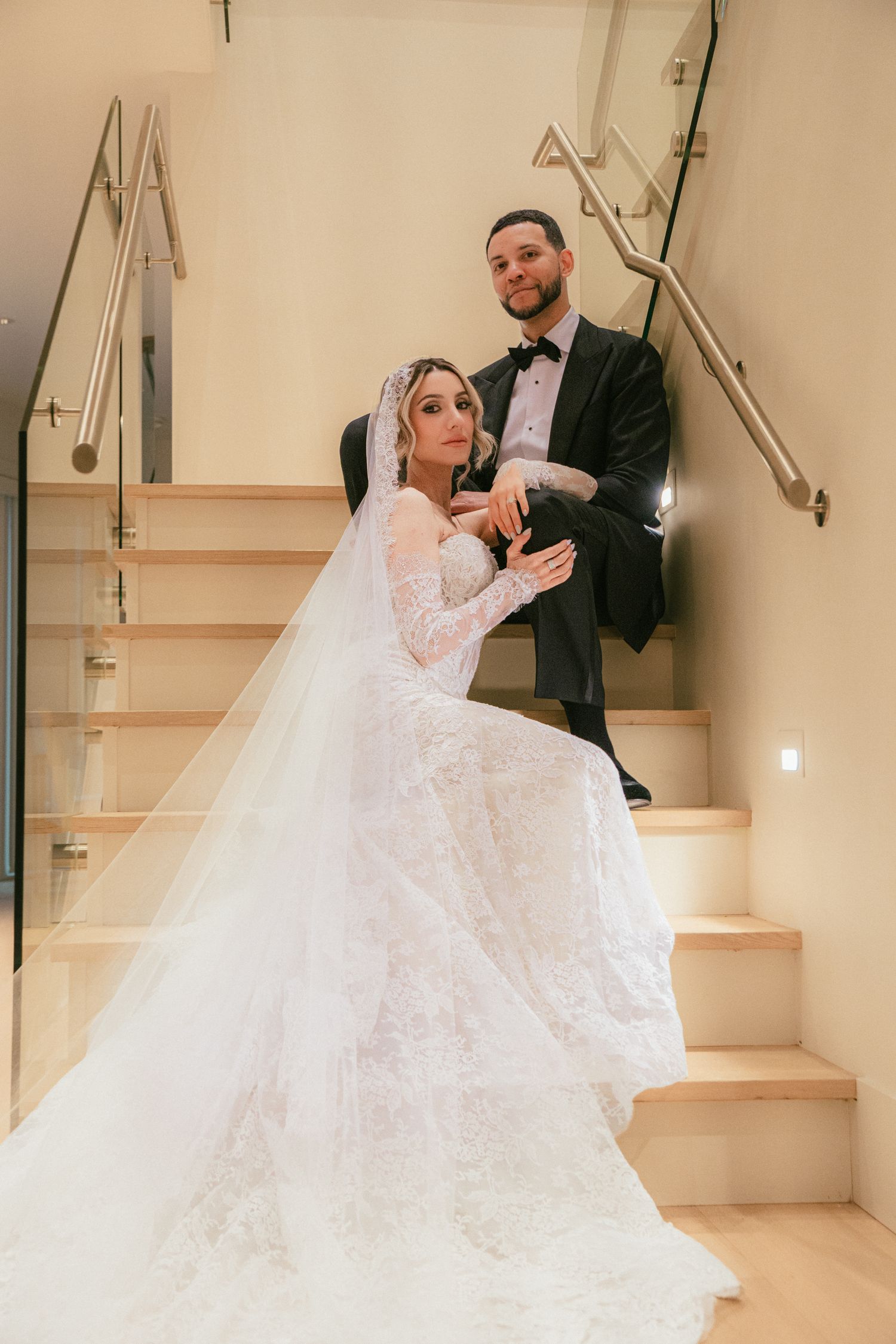 Bride in ballgown and veil poses with groom on modern staircase indoors.
