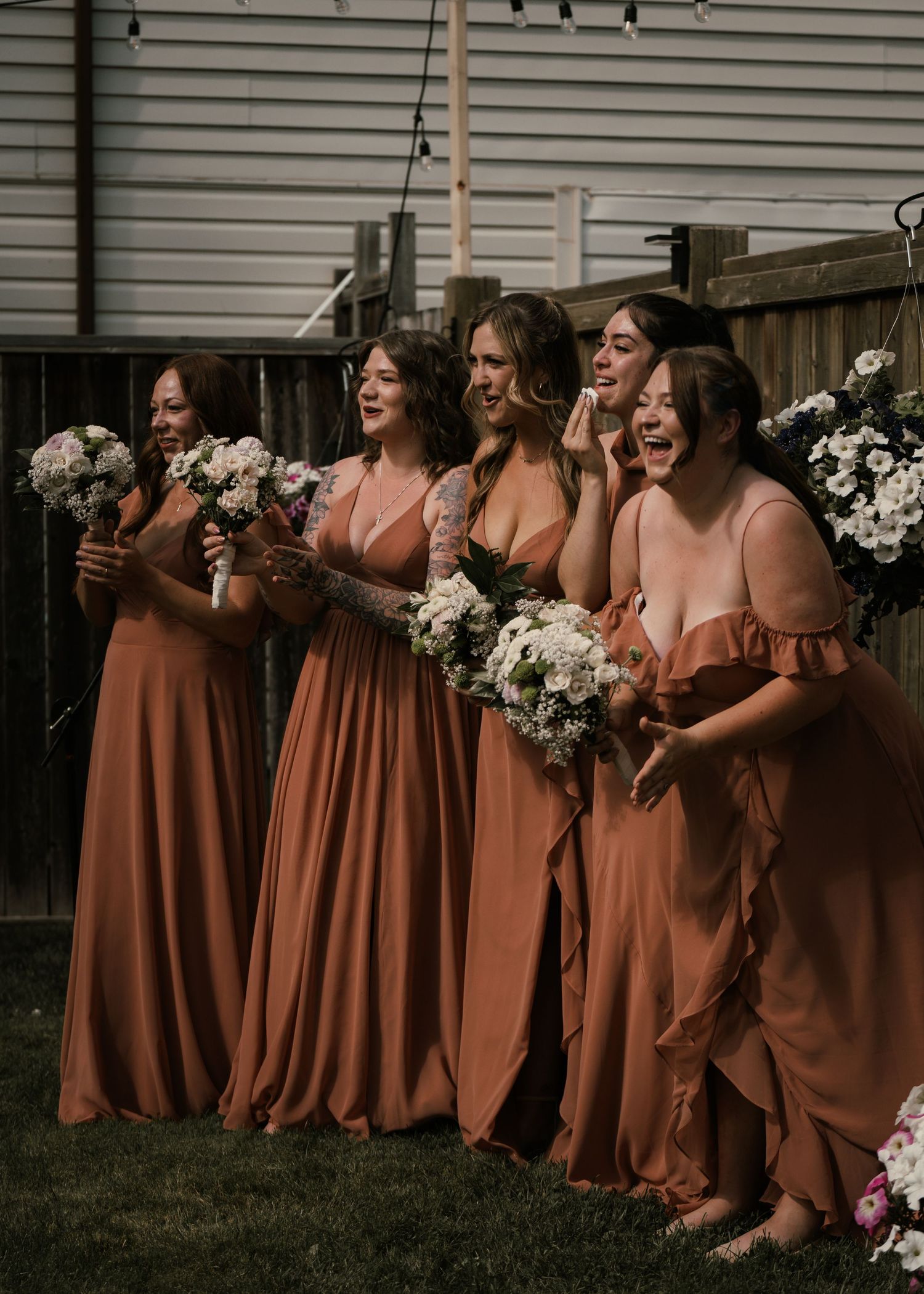 Five bridesmaids in rust-colored gowns holding white bouquets laugh joyfully outdoors at a backyard wedding ceremony.