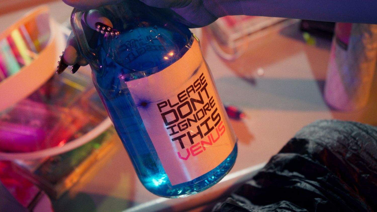 Blue liquid bottle with label reading Please Don't Ignore This Venus under neon pink lighting.