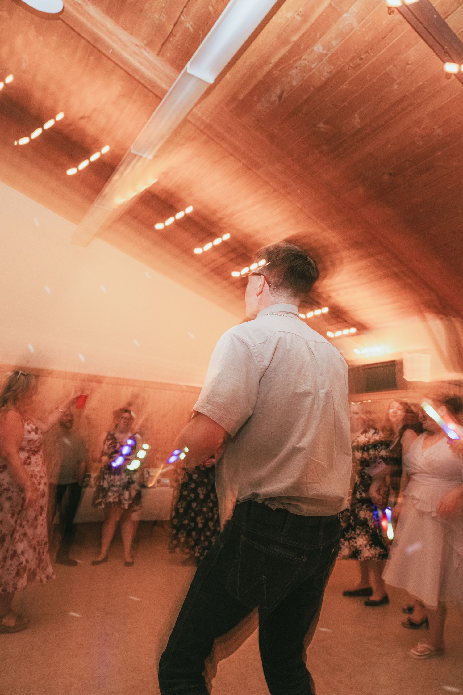 Guests dancing at a wedding reception in a warmly lit wooden venue with colorful LED lights.