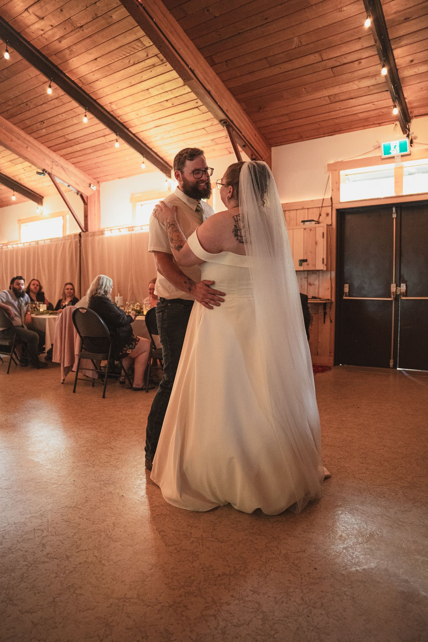 Newlywed couple shares a romantic first dance in a warmly lit barn reception hall with string lights.